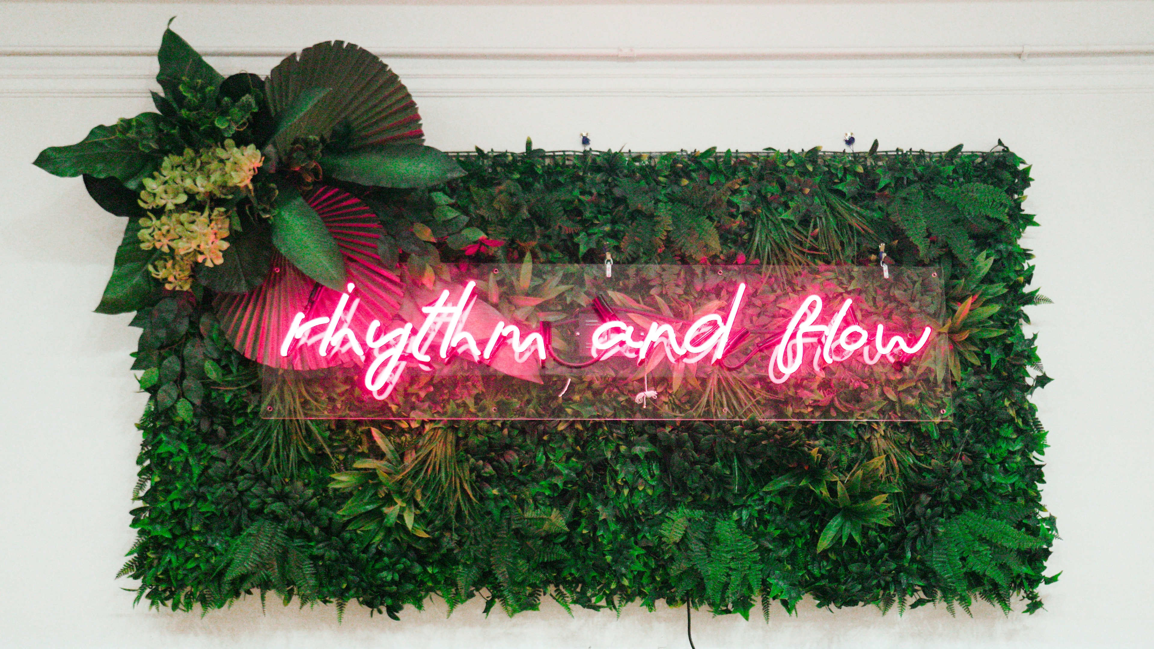 A neon sign reading "rhythm and flow" is mounted on a wall covered with various green plants.