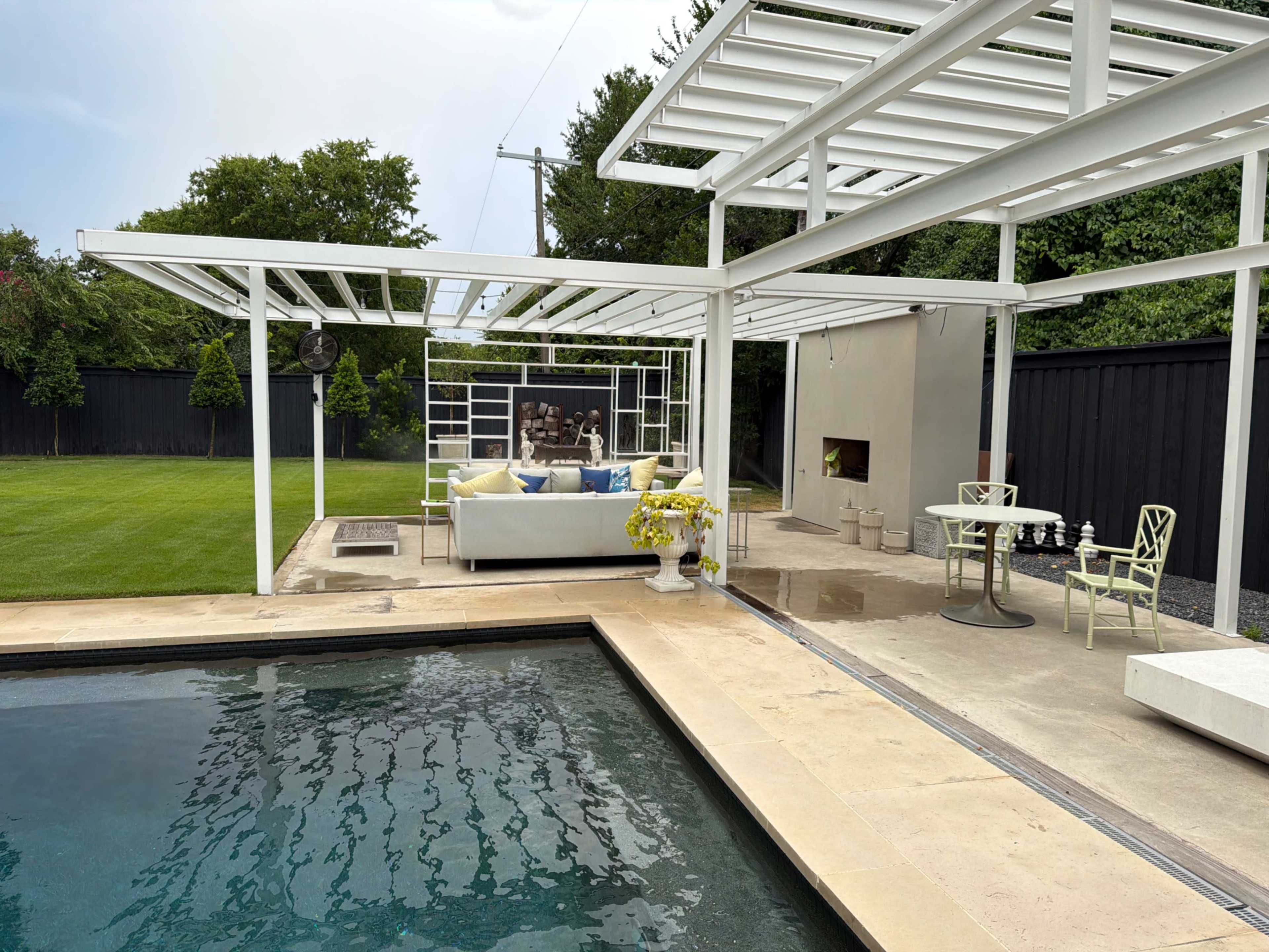 The image shows a modern outdoor area featuring a swimming pool, a covered seating area with a couch, and a stone patio adjacent to a lawn.