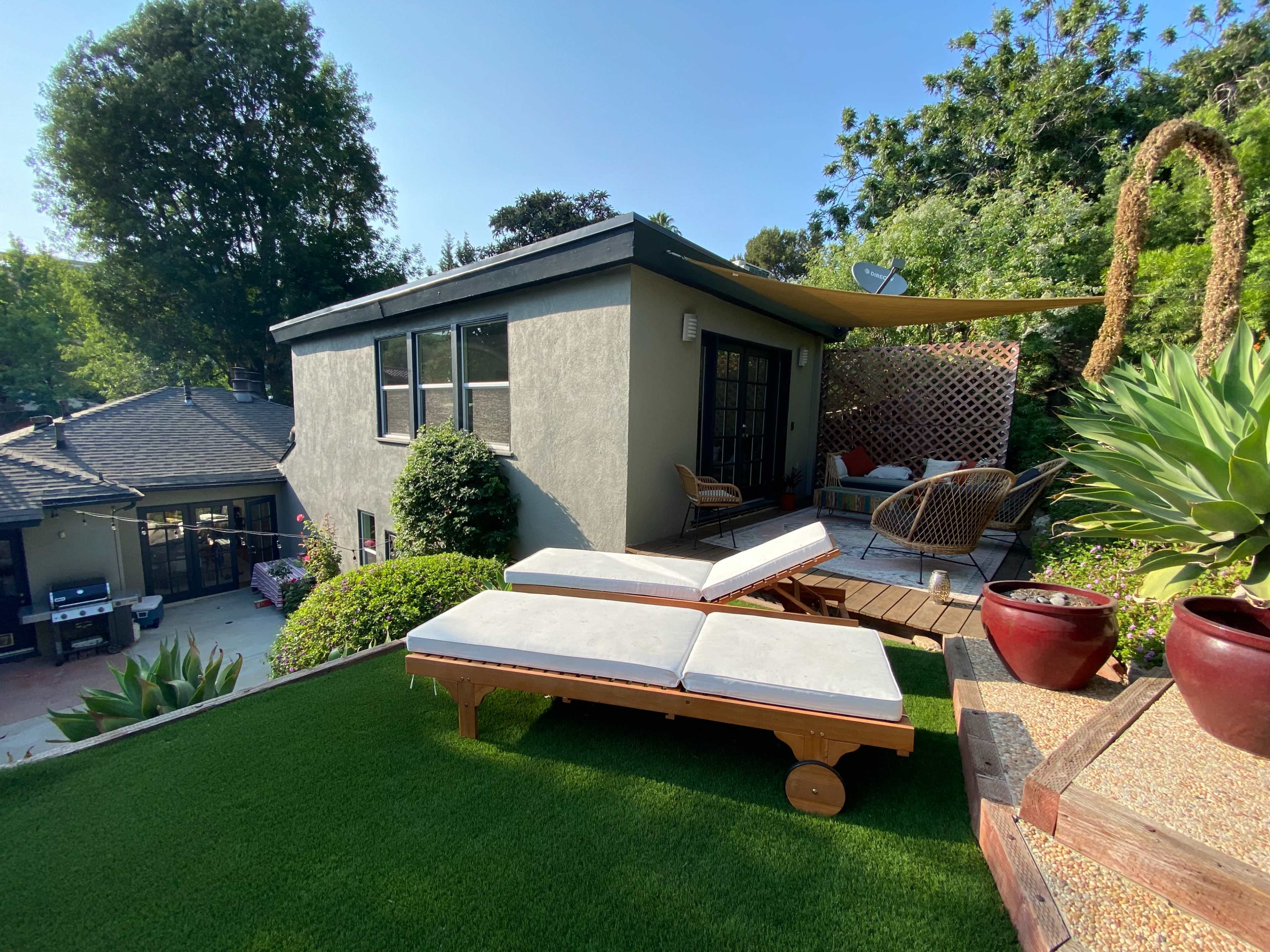 The image shows a backyard with two lounge chairs on artificial grass, a small patio area with seating, and a house surrounded by greenery.