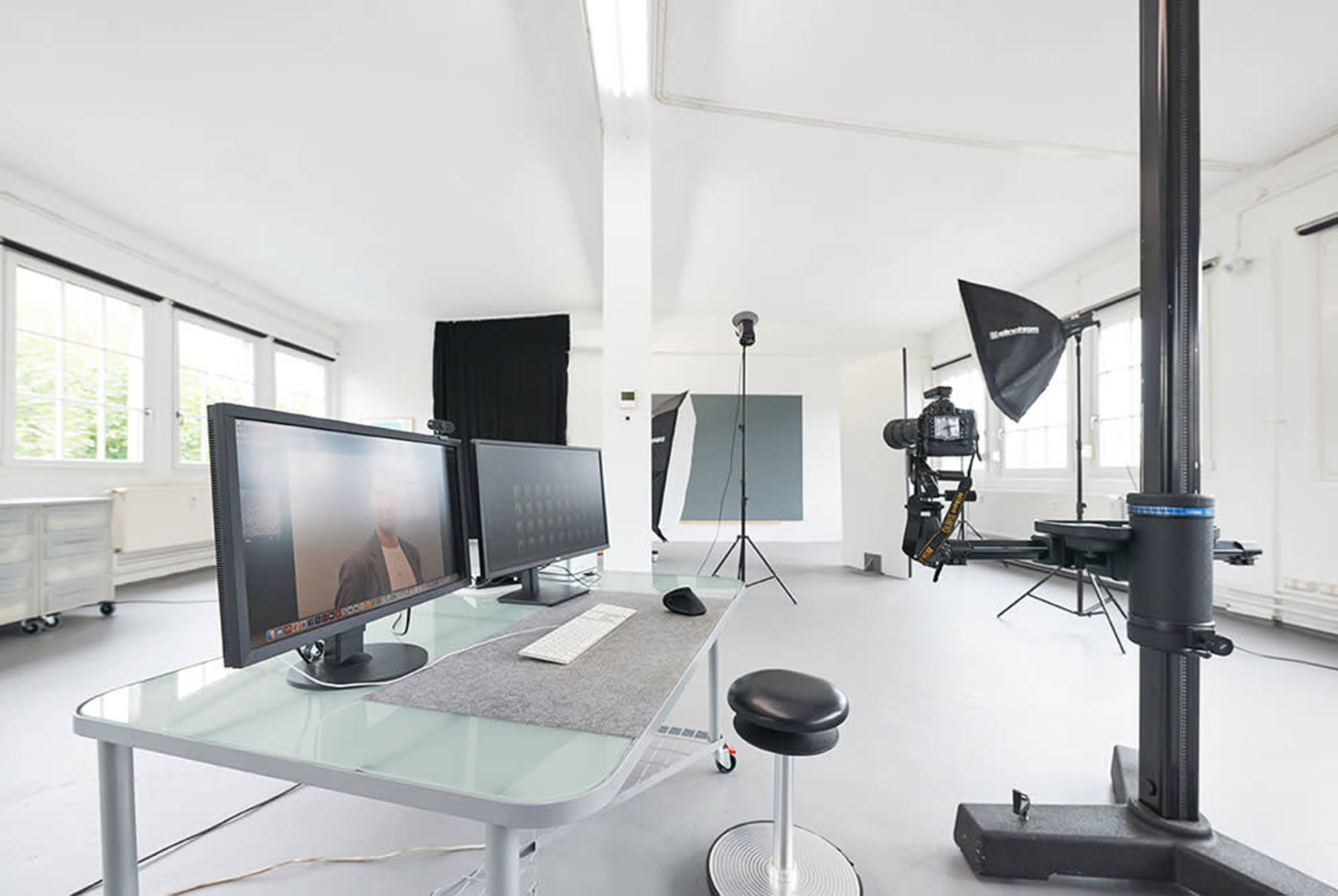 The image shows a modern photography studio equipped with a computer workstation, studio lighting, and a backdrop setup.