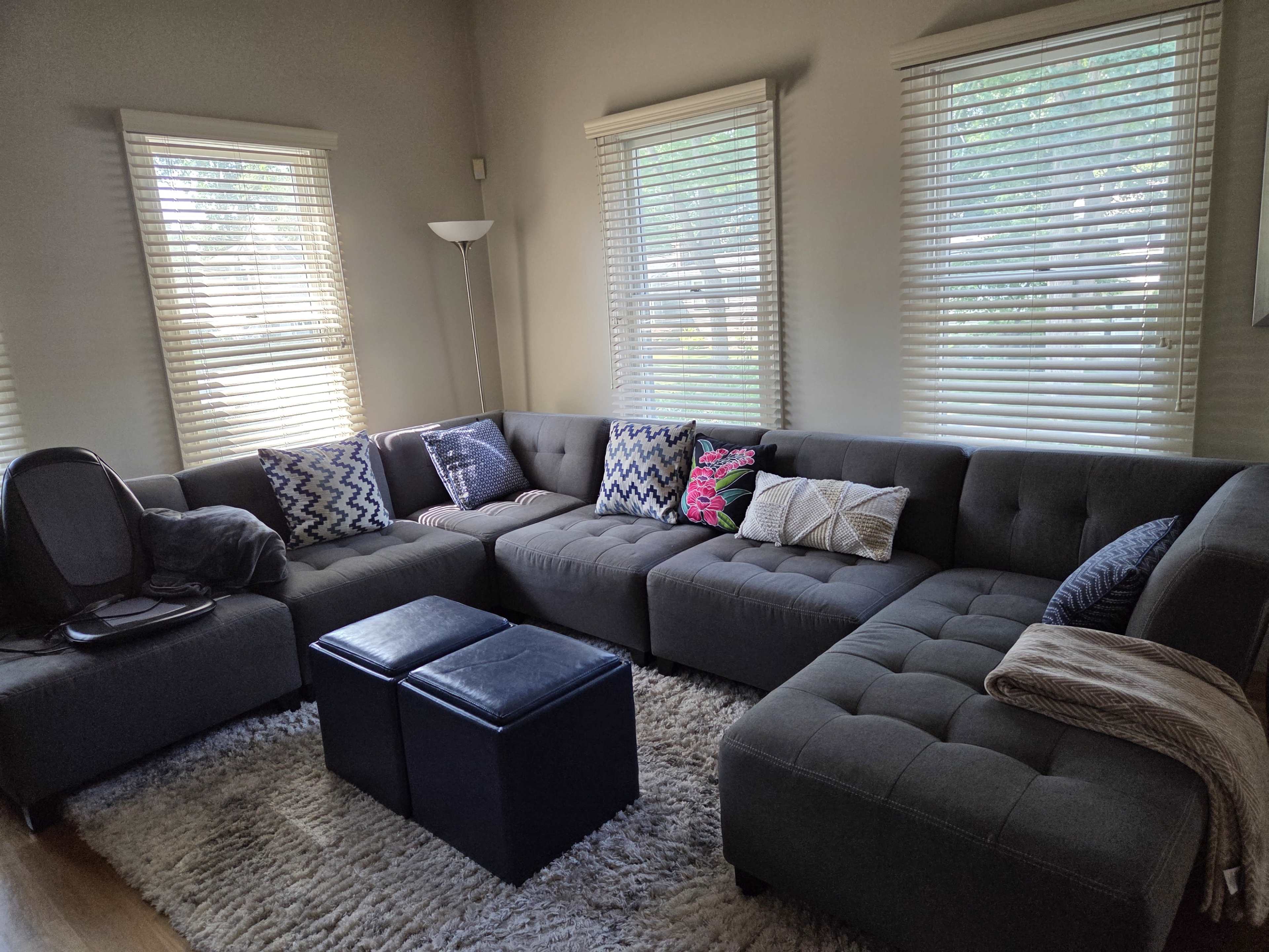 The image shows a gray sectional couch with decorative pillows arranged in a living room that has two windows with white blinds and a light rug on the floor.