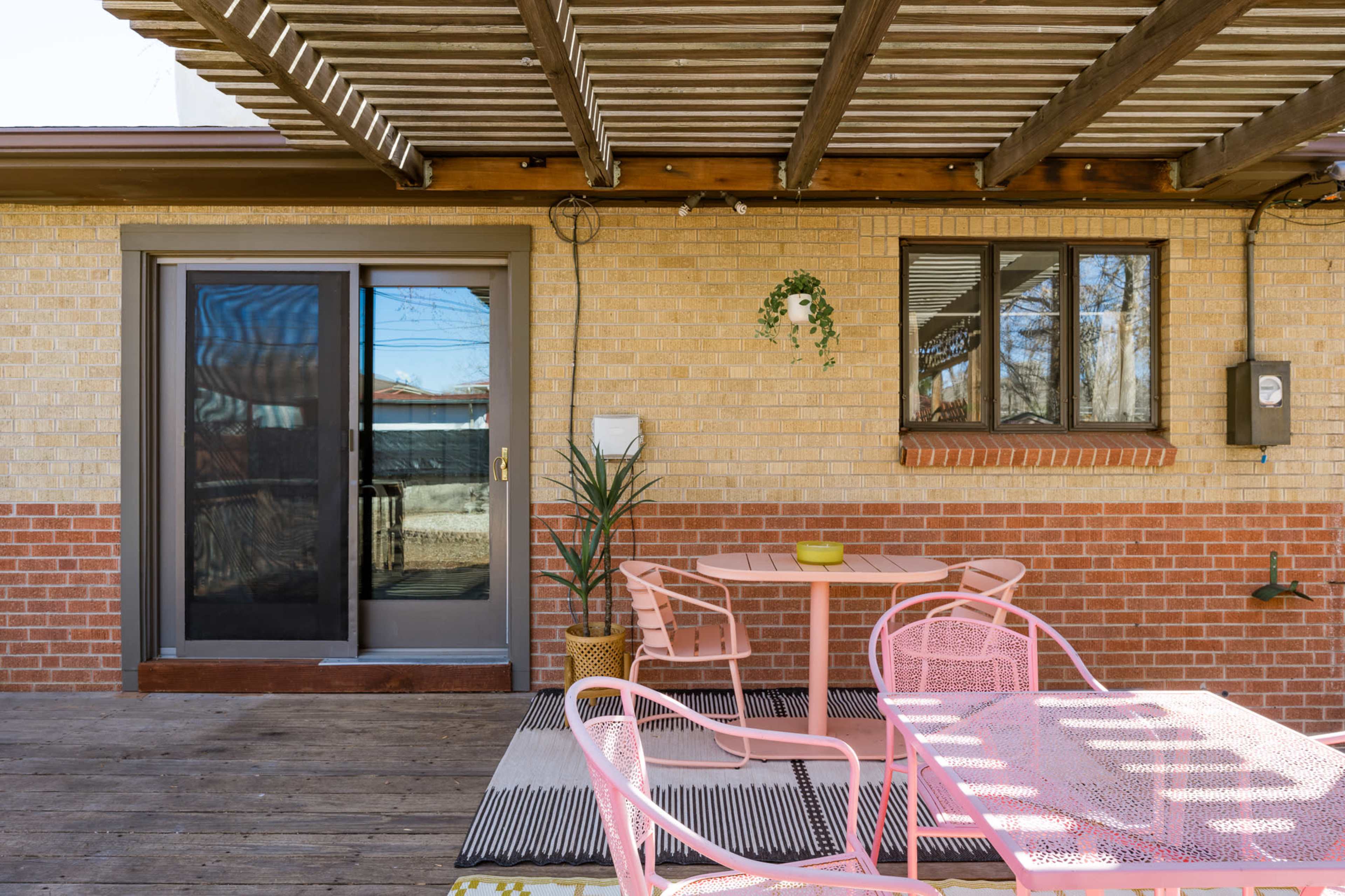 The image shows a patio area with a pink metal table and chairs, a potted plant, and sliding glass doors leading into a house.