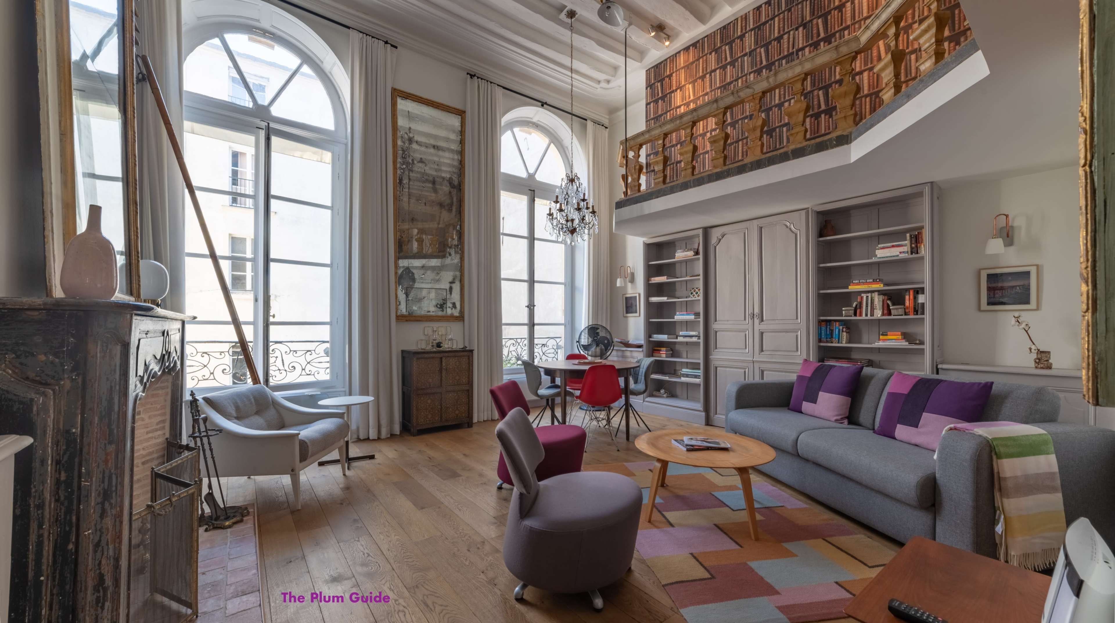 A modern living room with two large windows, a cozy seating area, and a lofted bookshelf overhead.