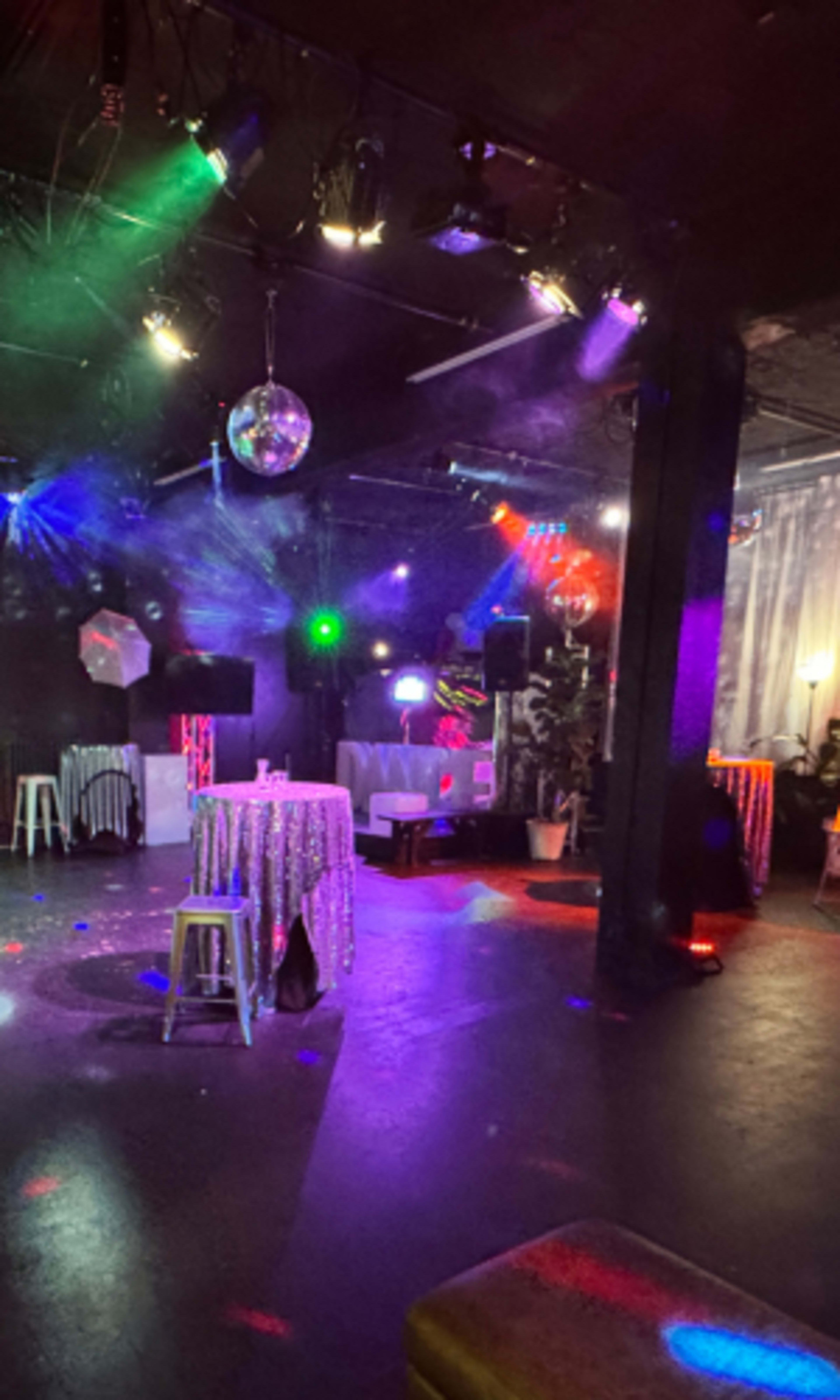 The image shows a dimly lit event space featuring colorful lights, a disco ball, and sparse seating arrangements with tables decorated with sparkly tablecloths.