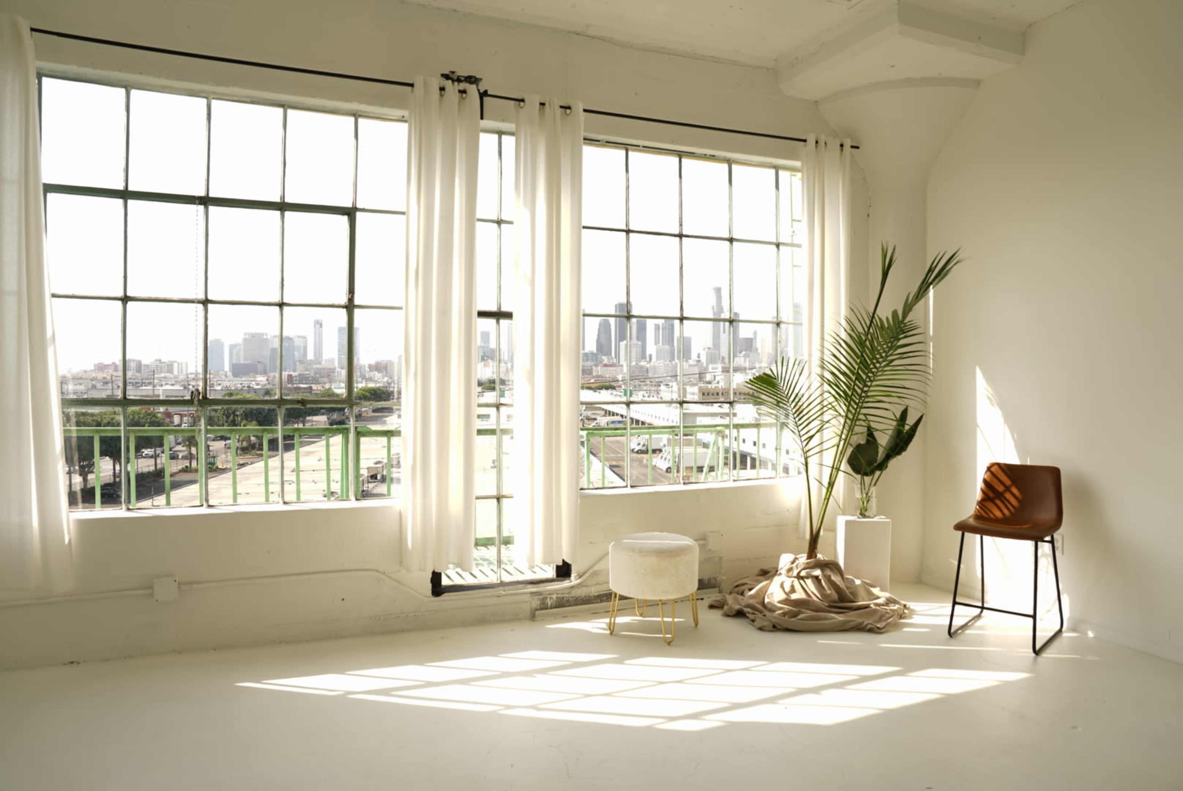A minimalist indoor space features large windows with city views, a potted plant, a chair, a small stool, and draped fabric on the floor.