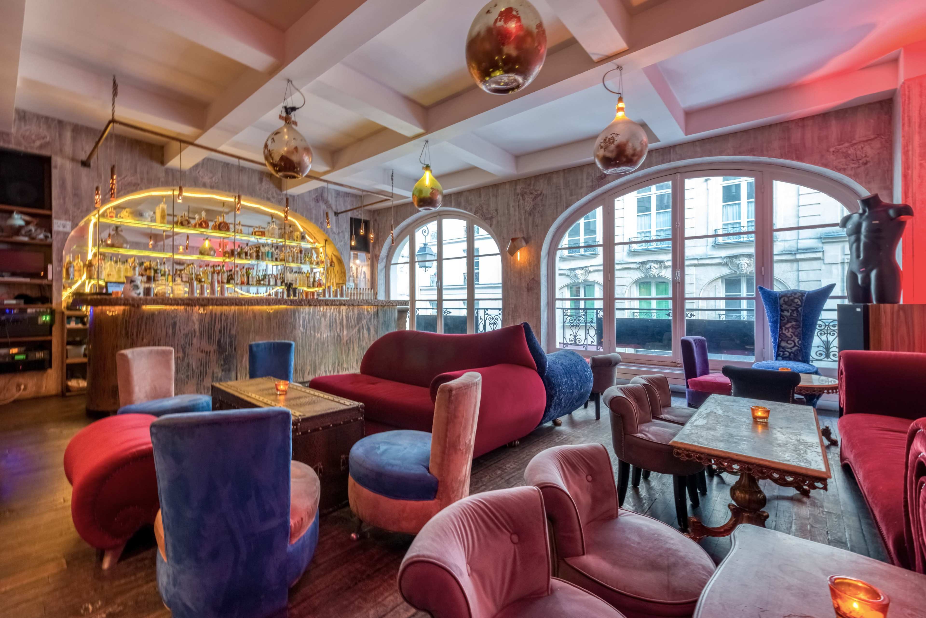 A stylish bar interior with plush seating, a circular bar backdrop, and large windows allowing natural light to enter the space.