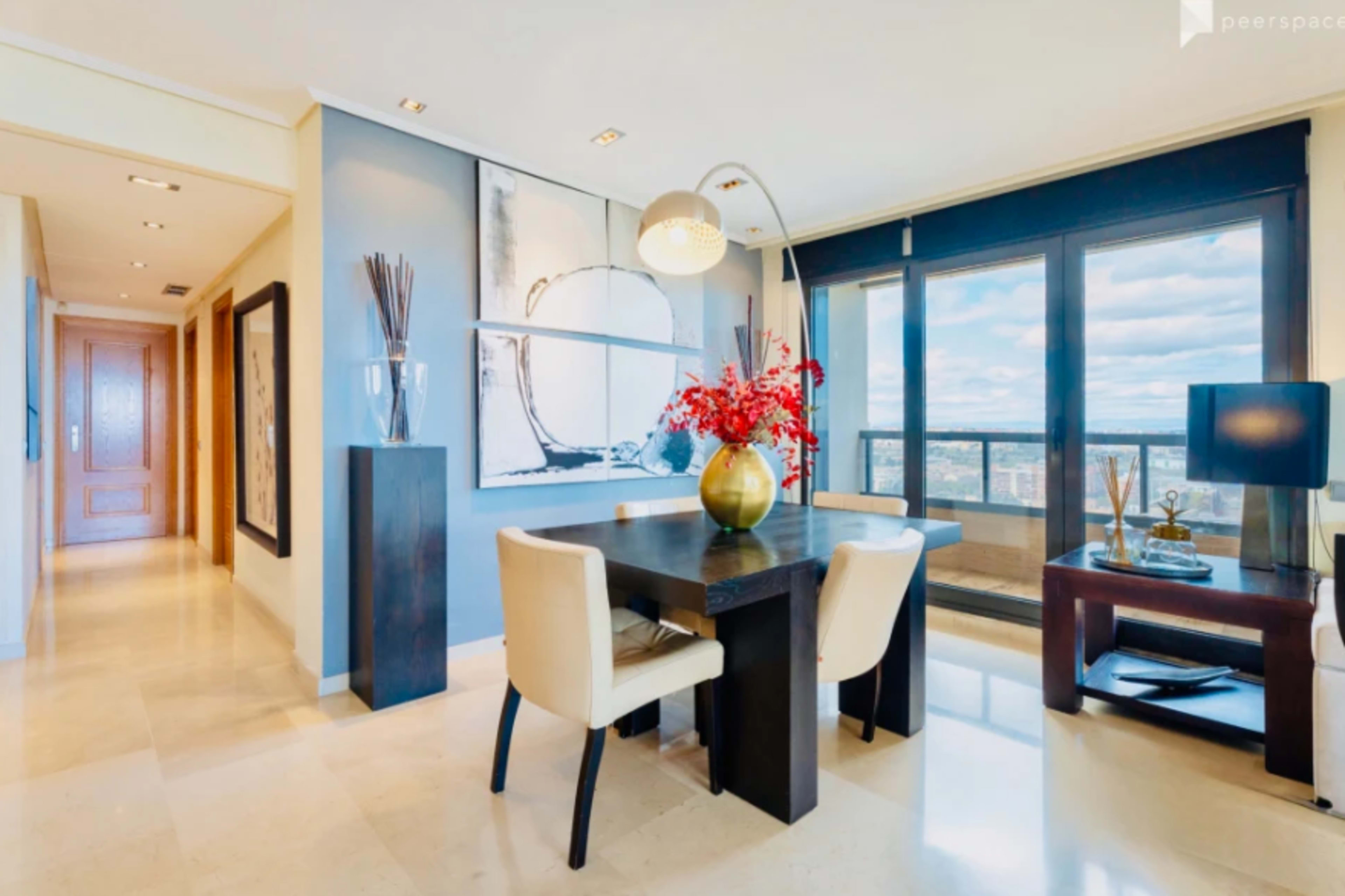 The image shows a modern dining area with a table, chairs, and decorative elements, adjacent to a hallway and large windows featuring a city view.