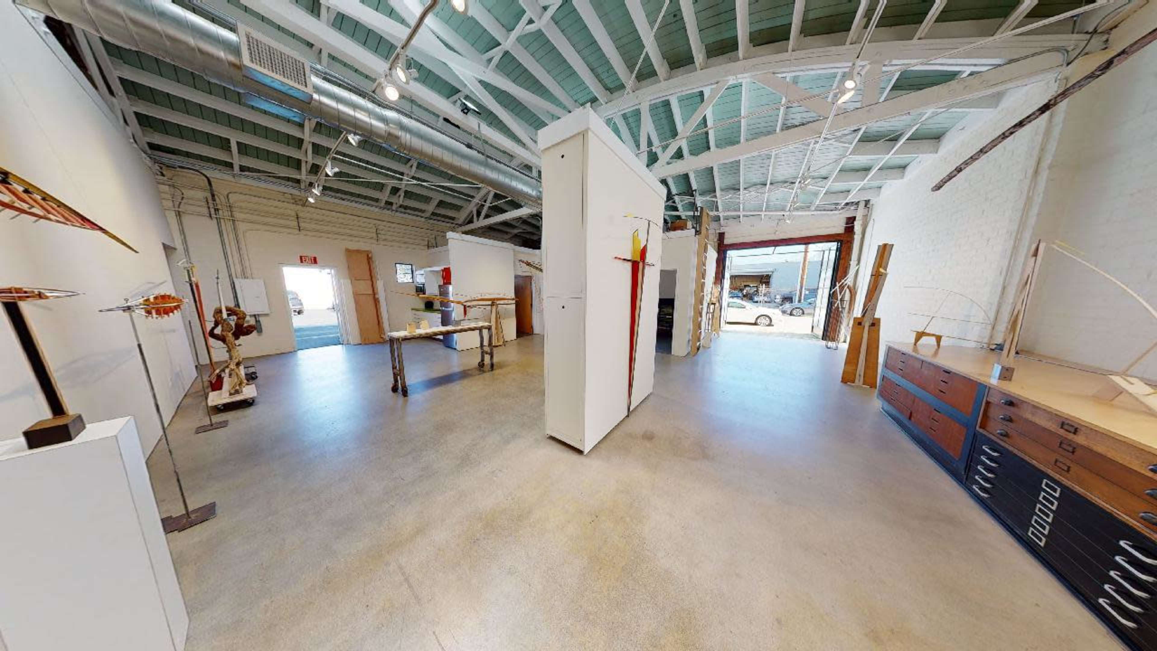 A spacious, well-lit art gallery with white walls and a concrete floor, featuring various sculptures and art installations.