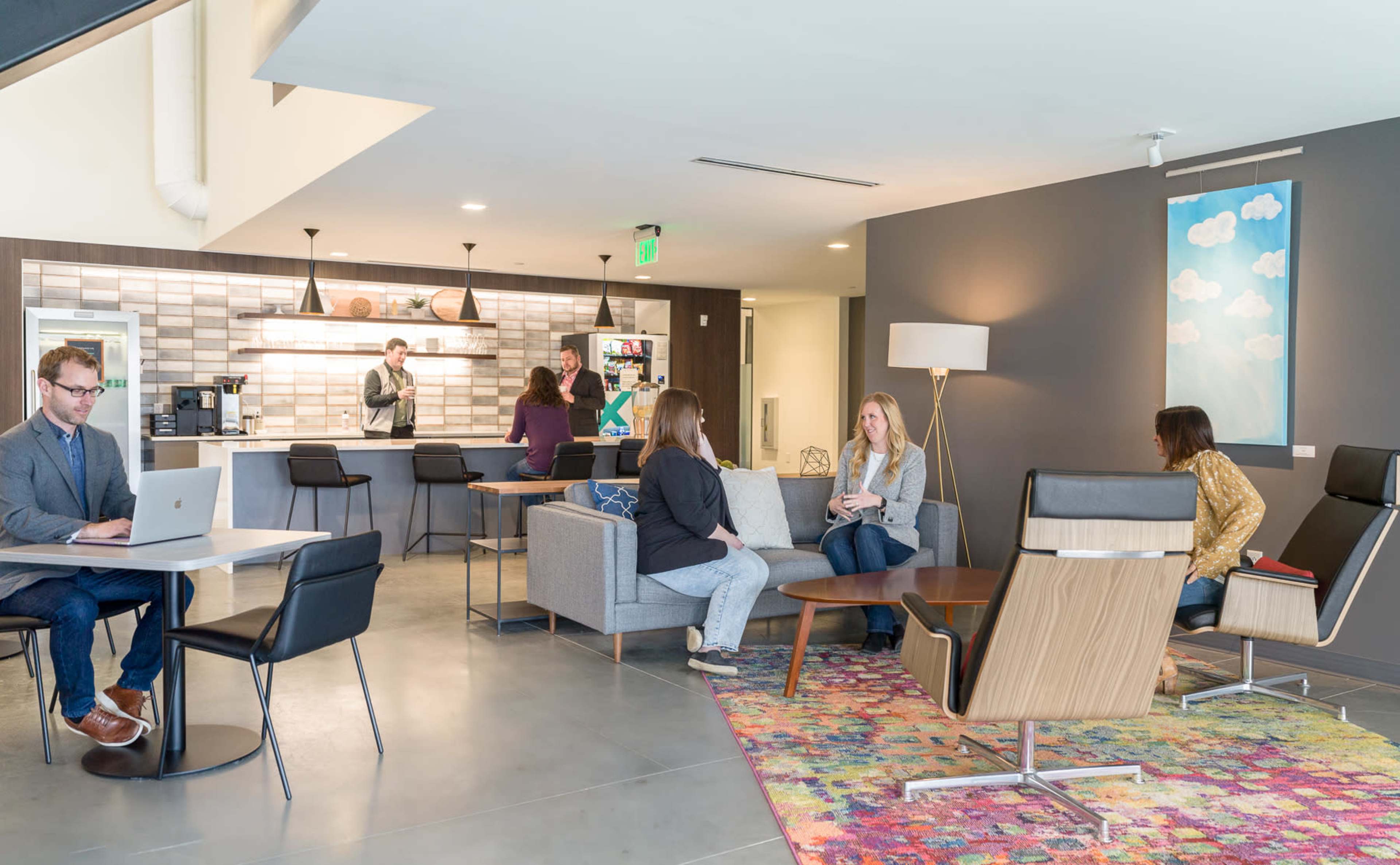 The image shows a modern co-working space with people engaged in various activities, including working on laptops and conversing.