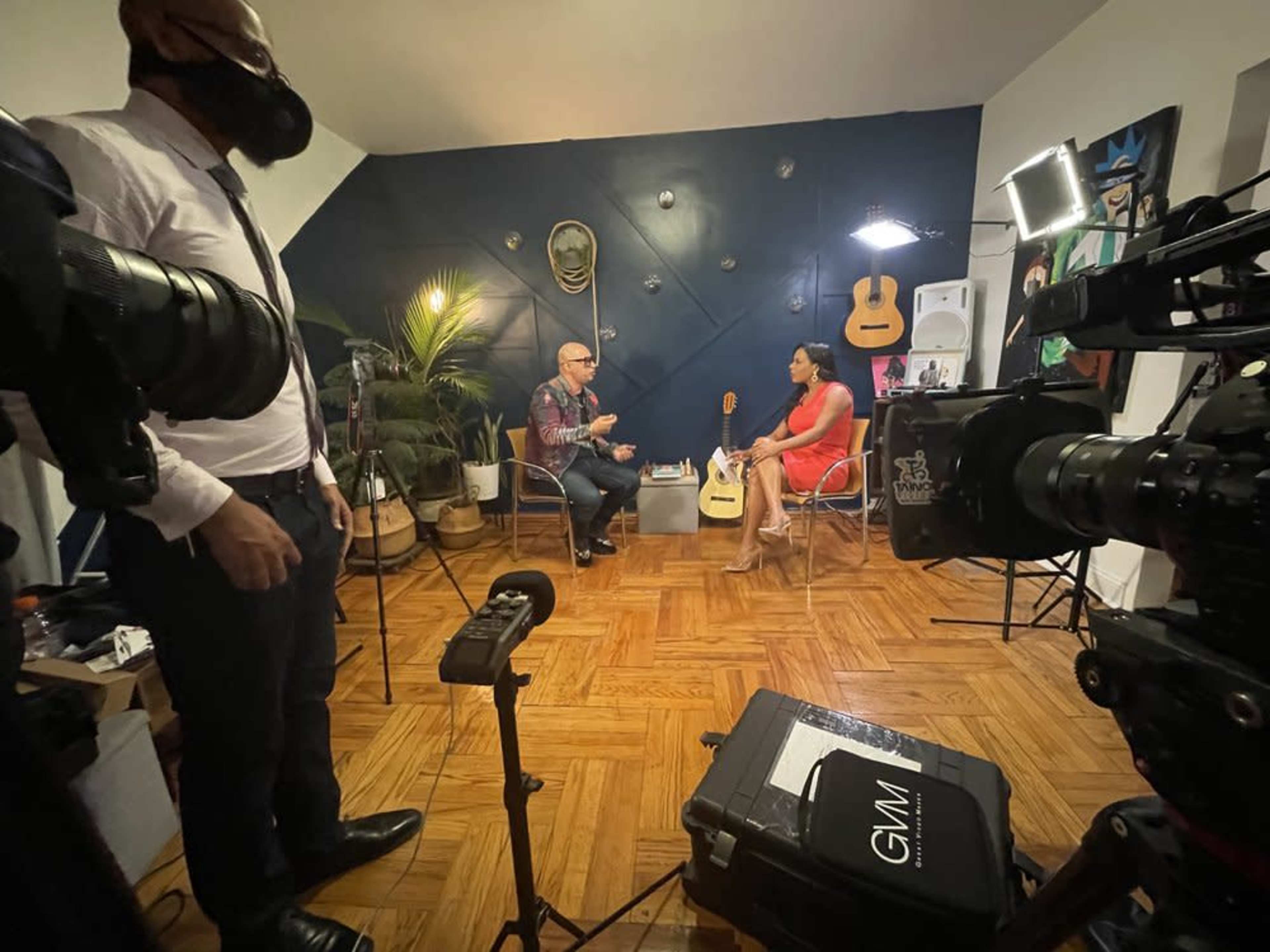 A man and a woman are seated and engaged in conversation in a well-decorated room equipped with cameras and lighting for a video shoot.