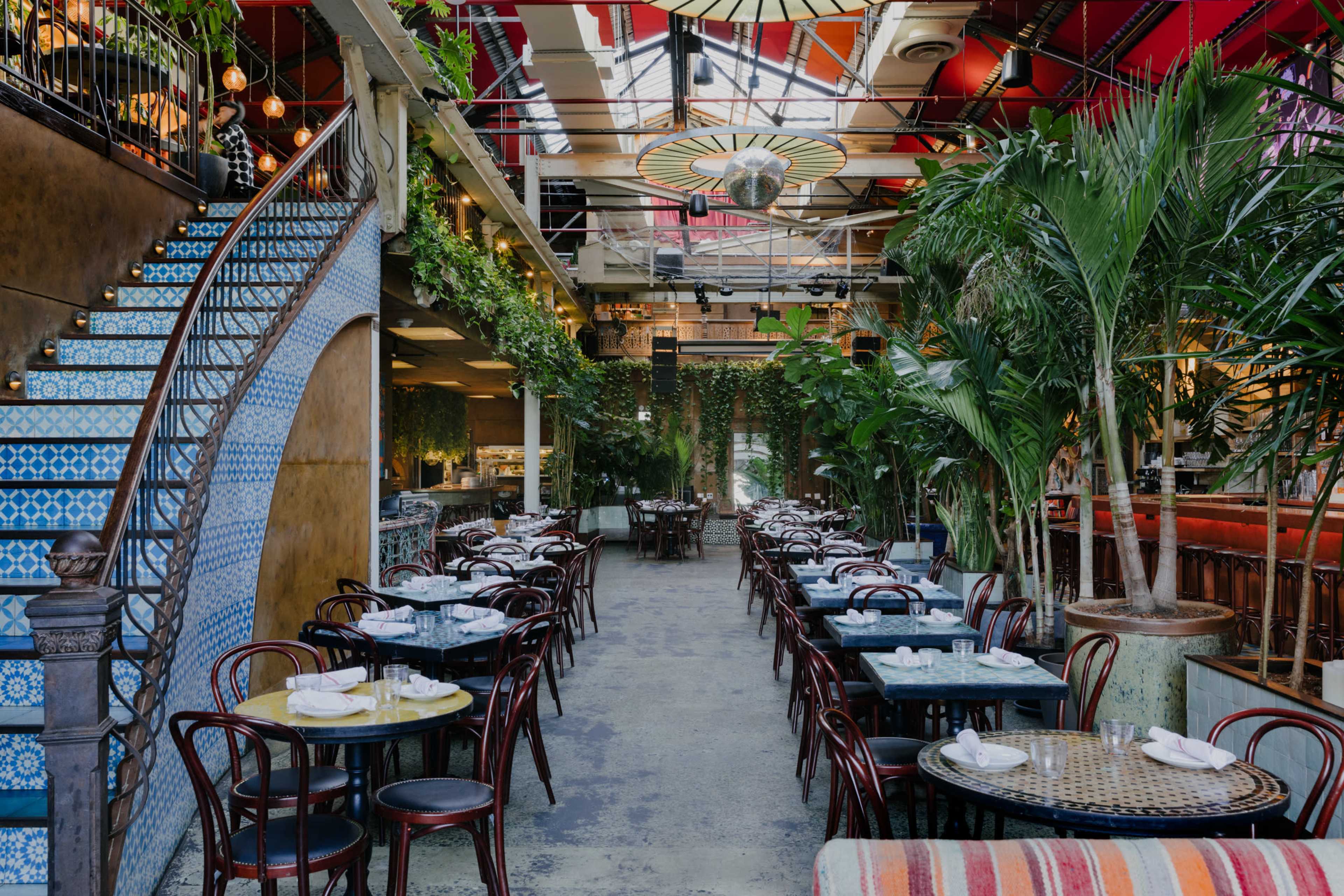 A spacious restaurant interior with circular tables, chairs, and decorative plants, featuring a staircase adorned with blue tiles.