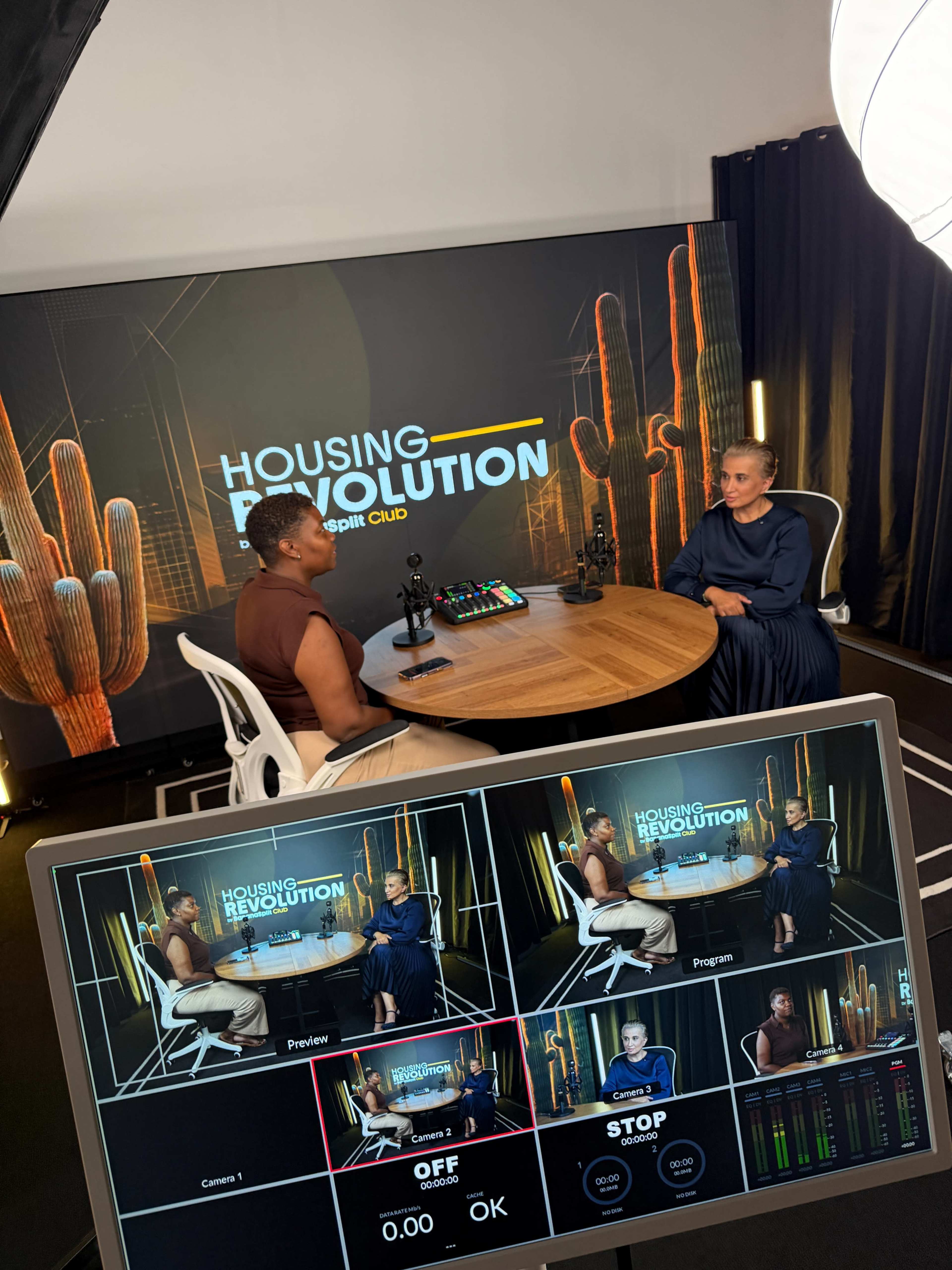 Two individuals sit at a round table in a studio setting, facing each other, with a large backdrop displaying "Housing Revolution" and various video production equipment visible in the foreground.