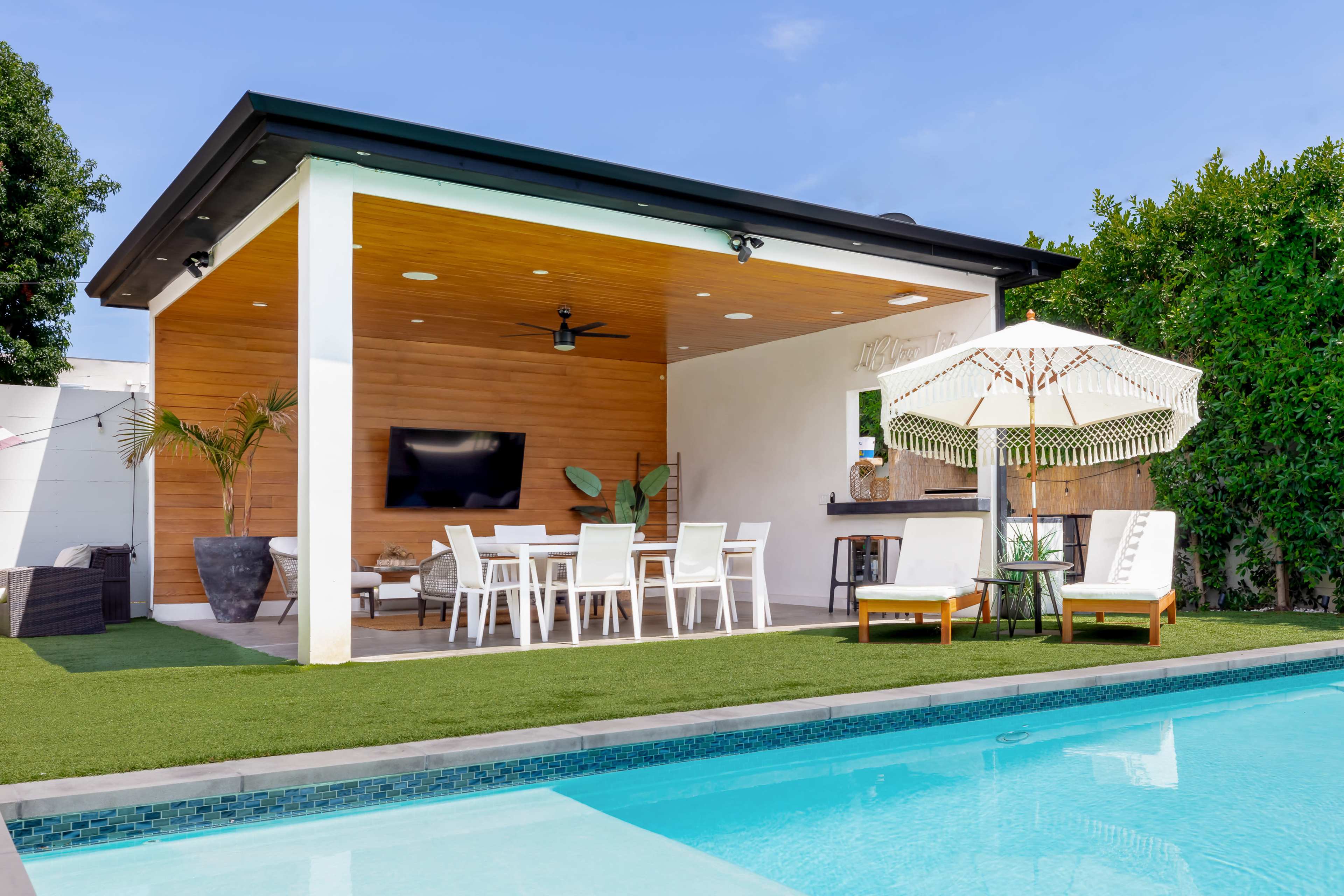 A modern outdoor living space with a covered patio, dining area, and a swimming pool surrounded by lush greenery.