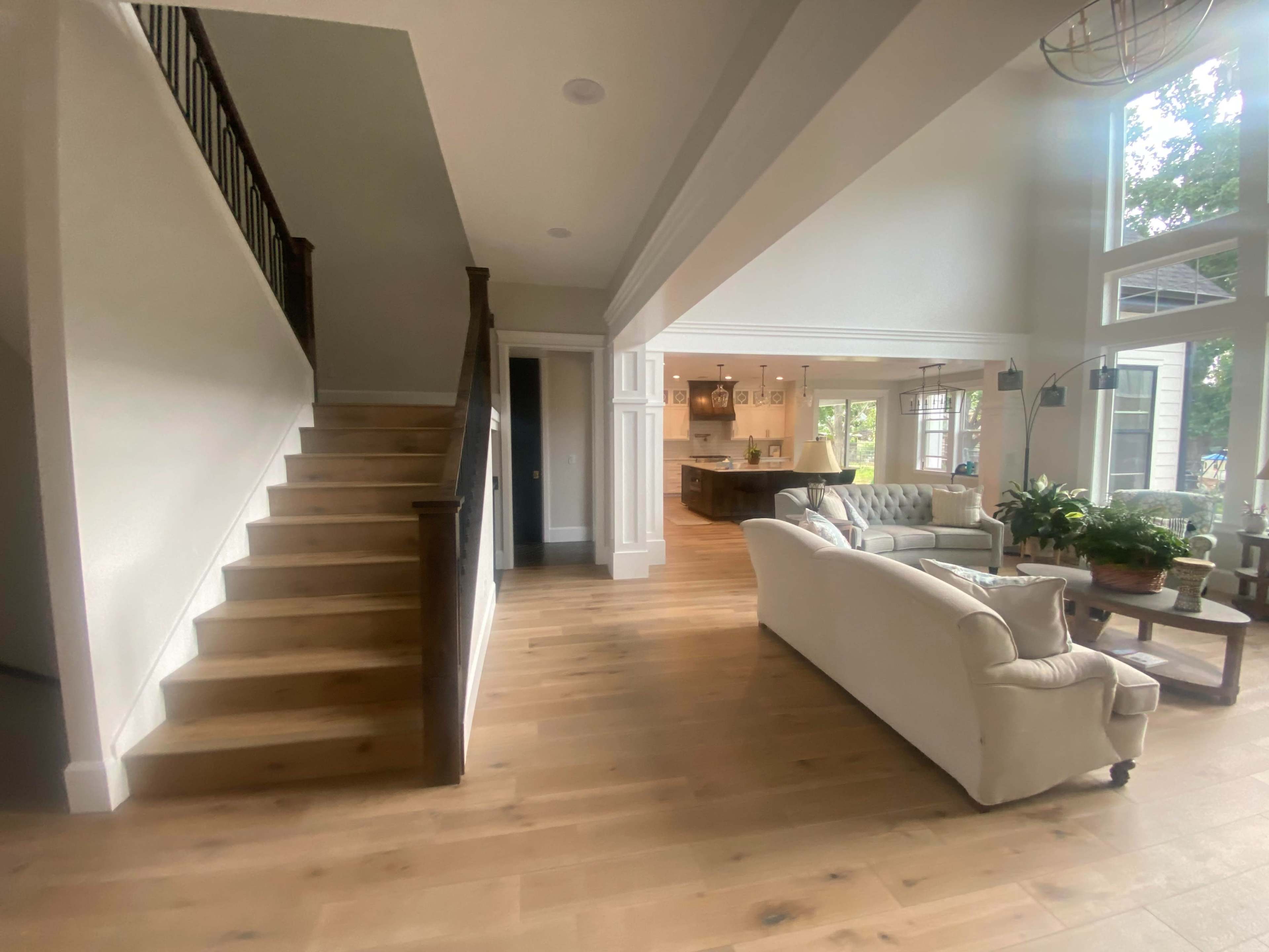 A bright, open living space featuring a staircase on the left, a sitting area with light-colored furniture, and a view into a kitchen area through an archway.