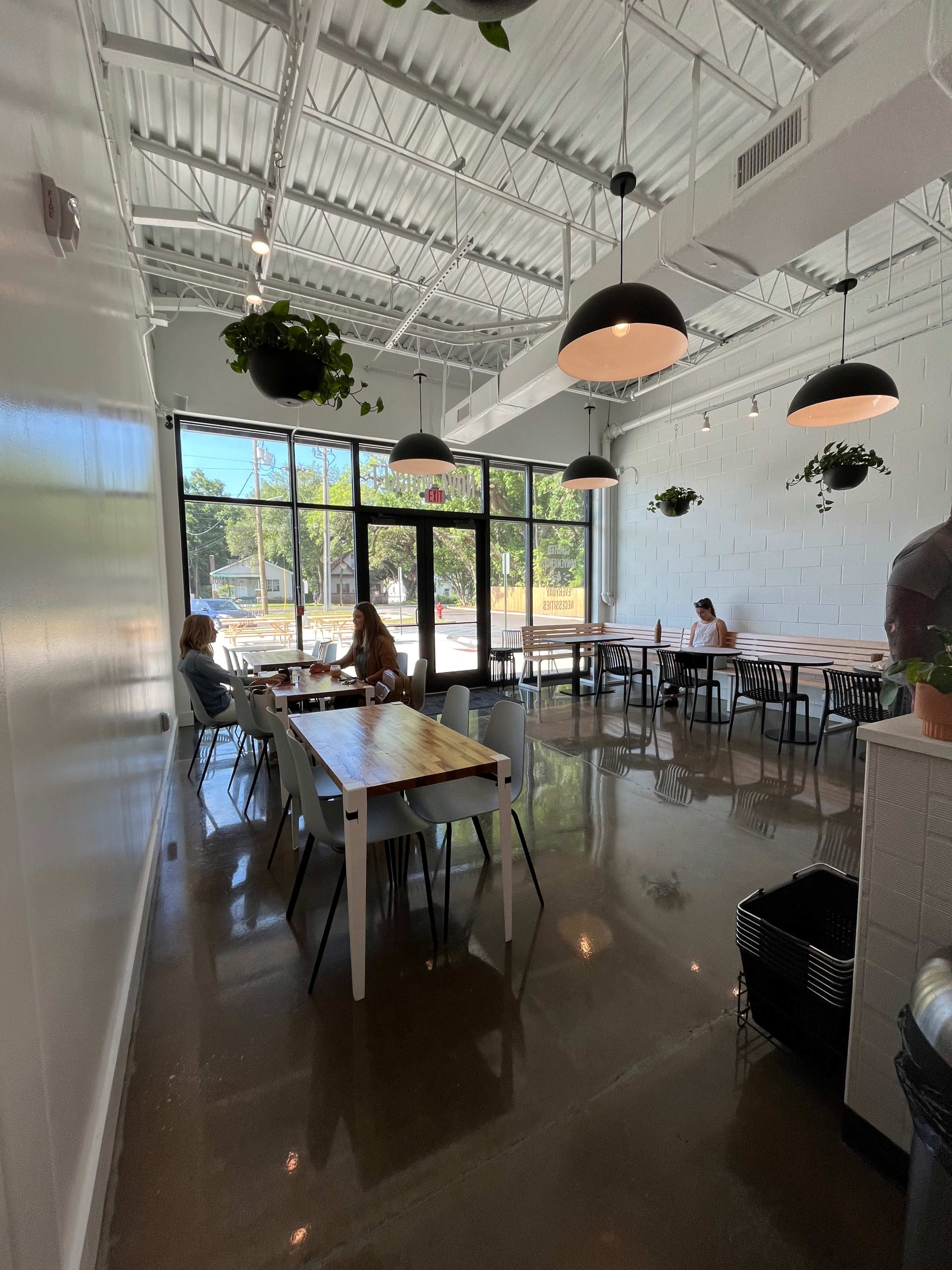 A bright, minimalist café with tables and chairs, featuring indoor plants and large windows overlooking the outside.