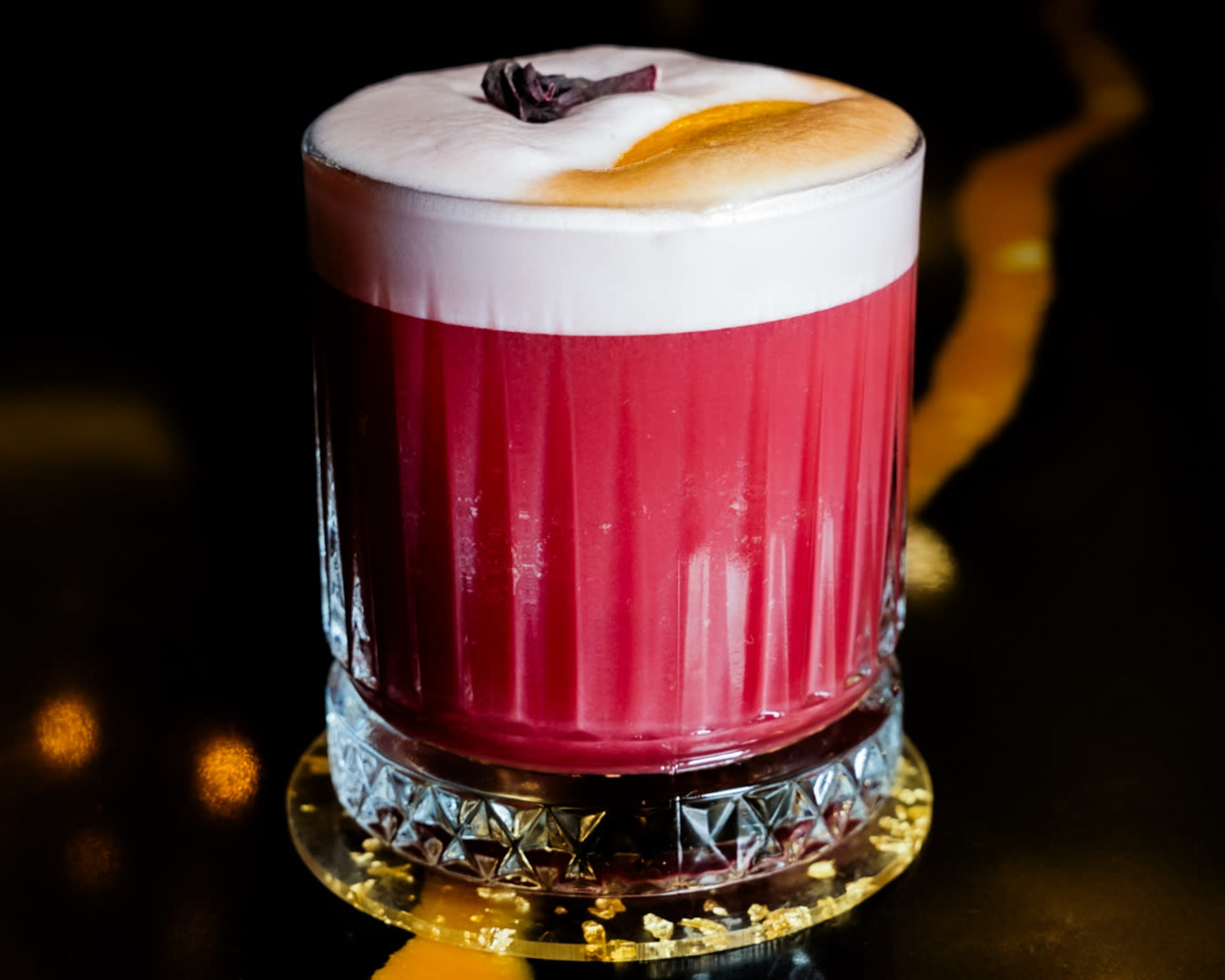 The image shows a cocktail in a crystal glass, featuring a vibrant red drink topped with a frothy layer and garnished with a piece of dried fruit.