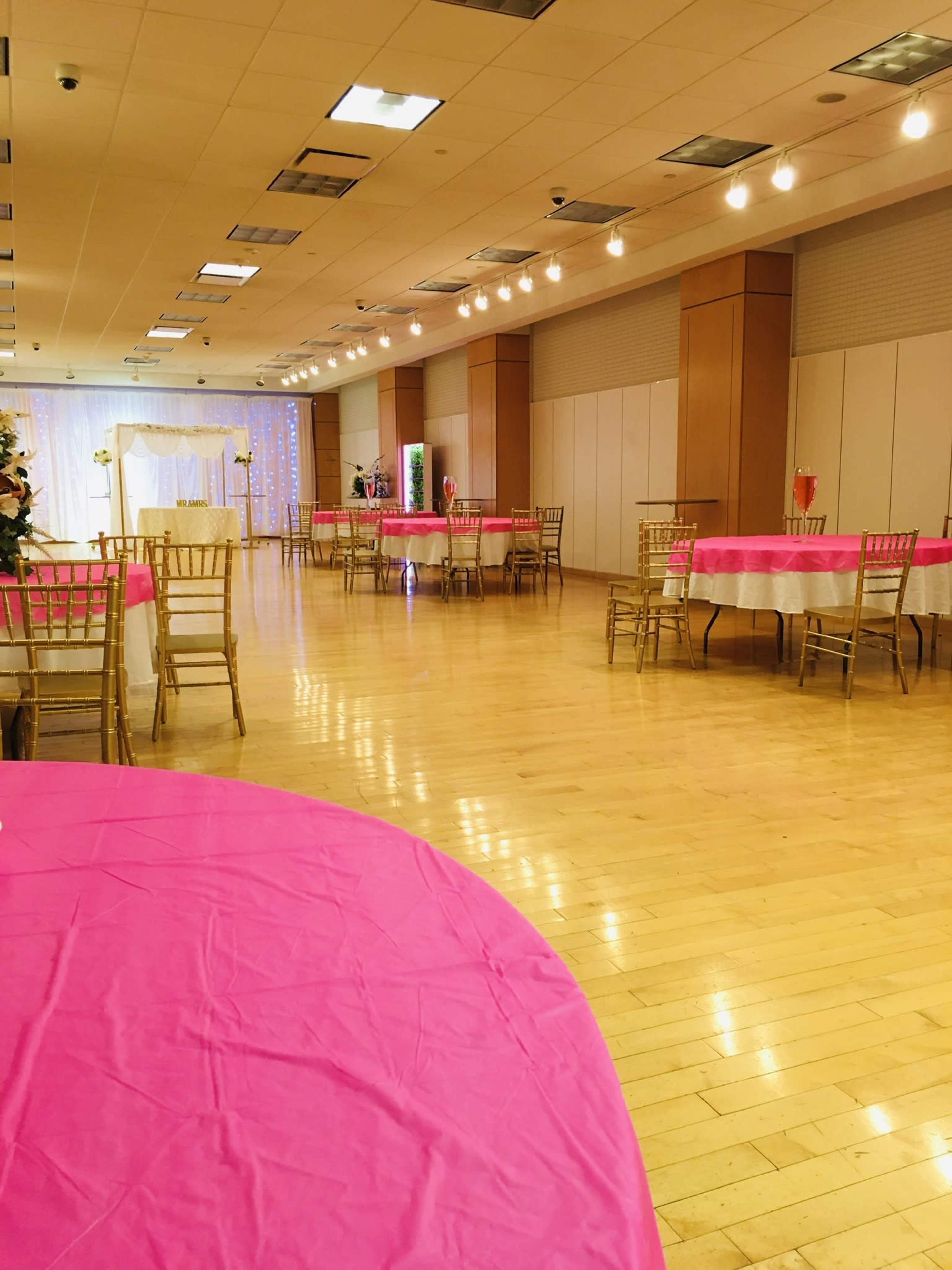 An elegantly arranged banquet hall with pink tablecloths covering several round tables and a decorative backdrop at one end of the room.