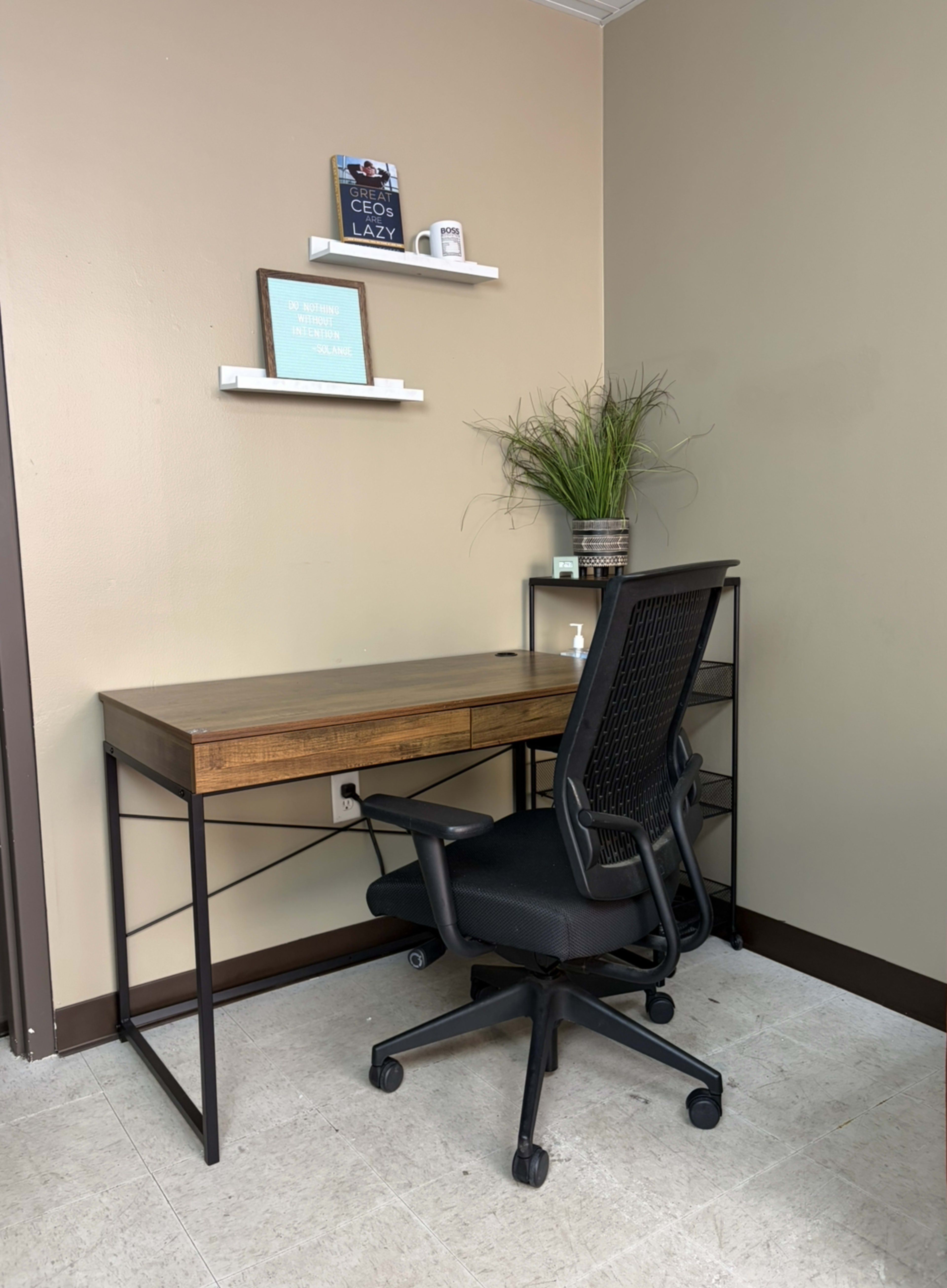The image shows a minimalist workspace featuring a wooden desk, a black office chair on wheels, and two shelves mounted on the wall with decor items.