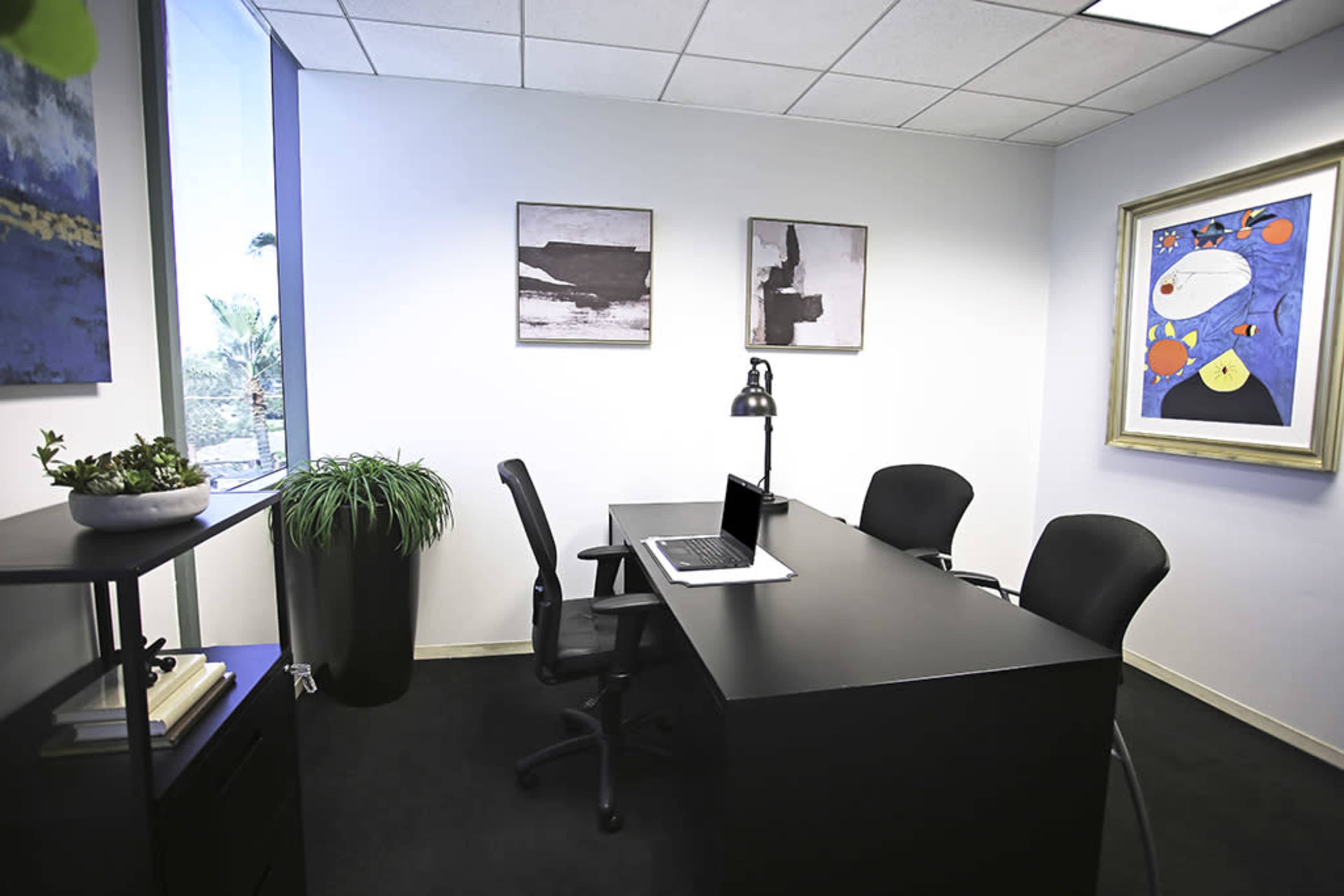 A modern office space with a black desk, two chairs, a laptop, and framed artworks on the walls.