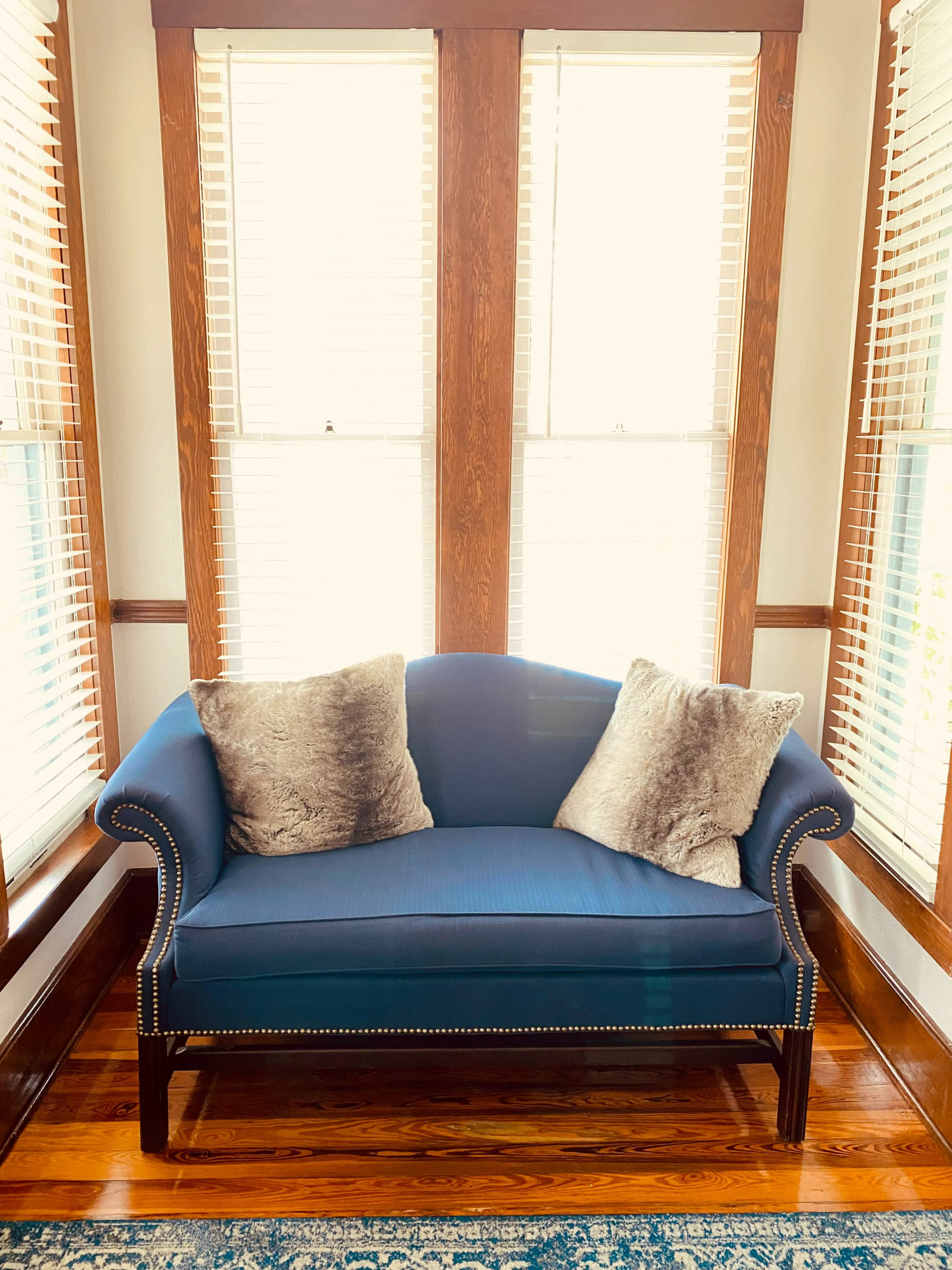 A blue loveseat with two decorative pillows sits in front of two windows with wooden blinds.