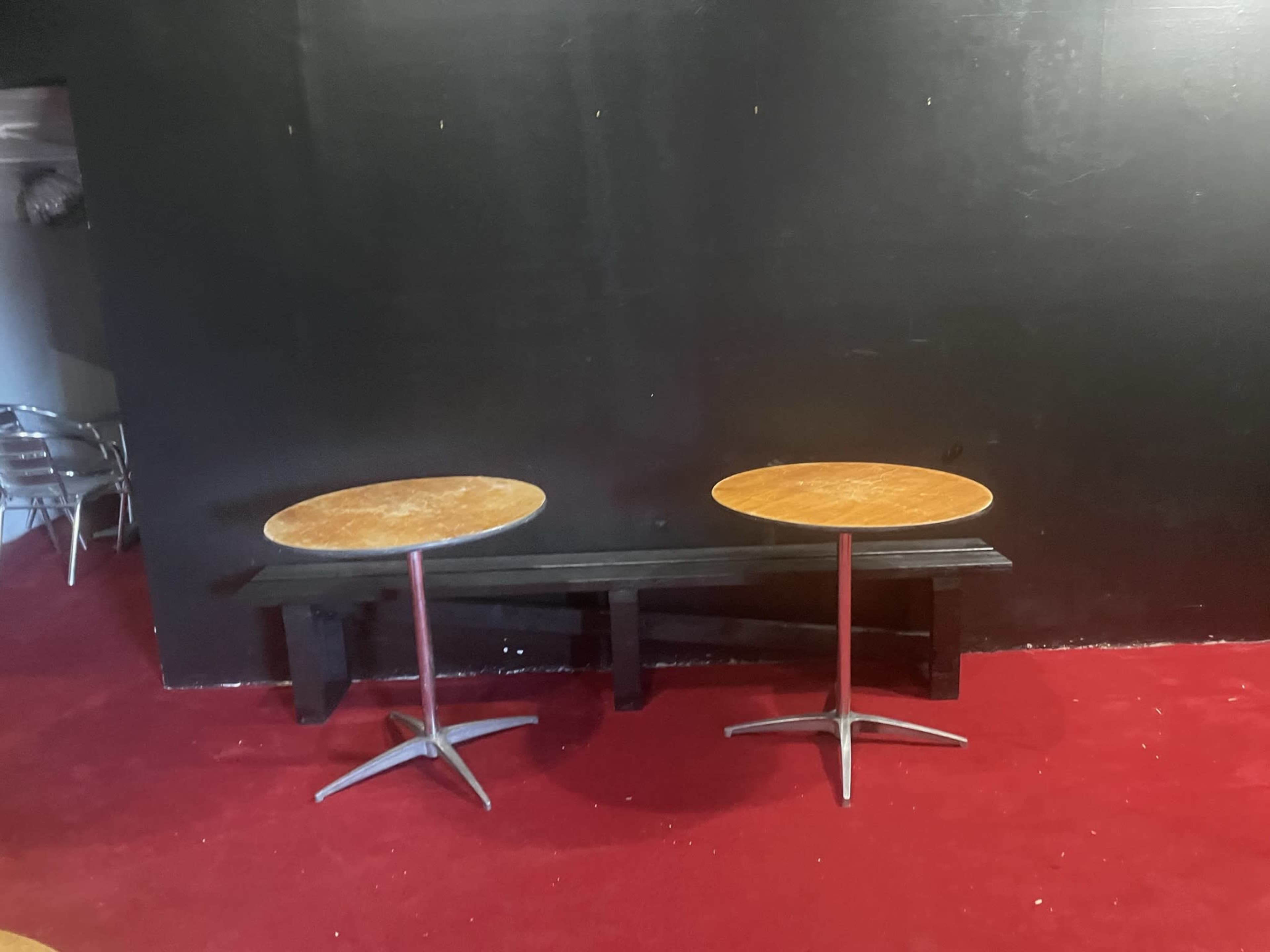 Two round wooden tables with metal bases are positioned on a red carpeted floor in front of a black wall.