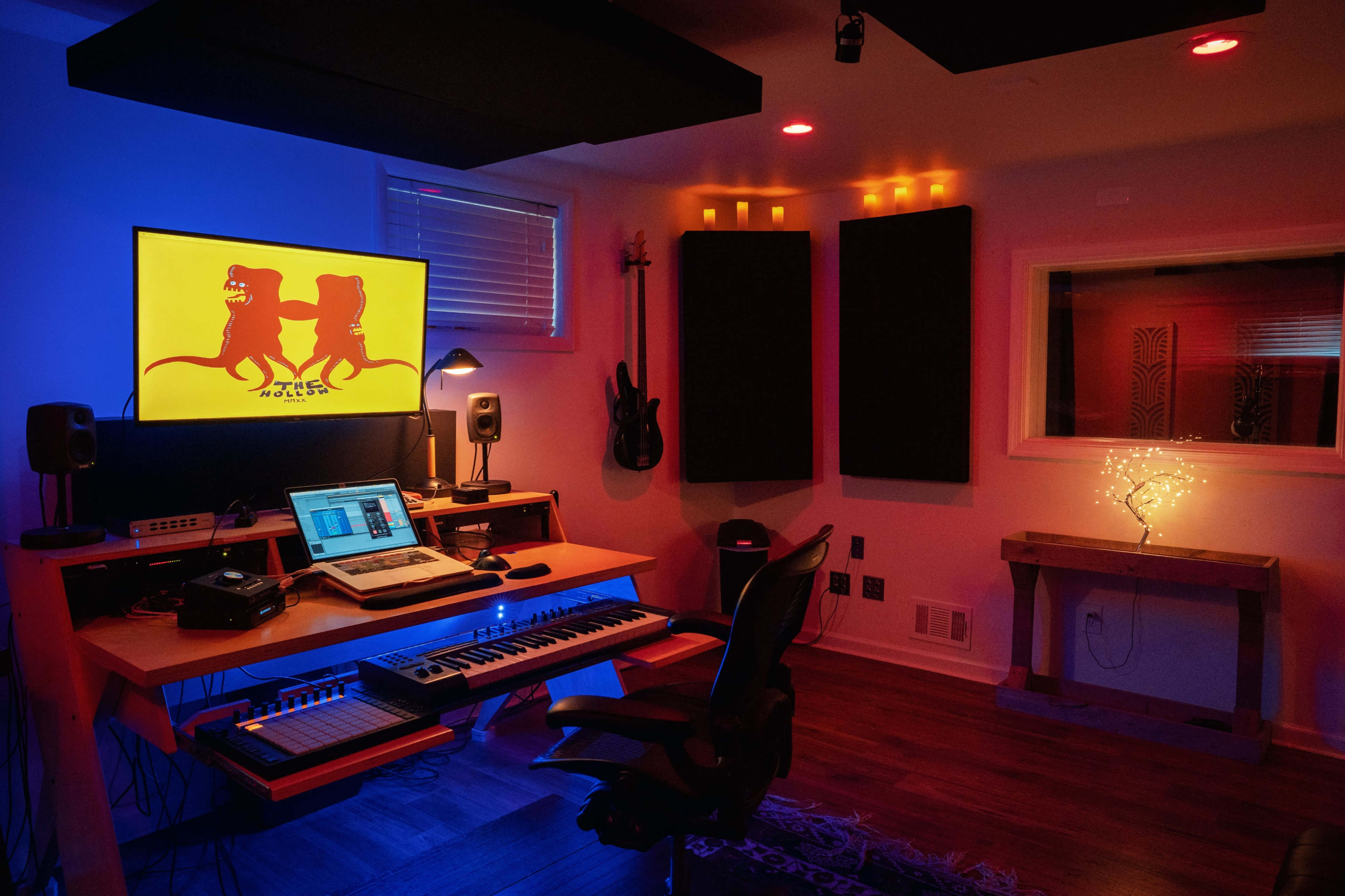 A dimly lit recording studio with a computer and MIDI keyboard on a wooden desk, surrounded by soundproof panels and colorful ambient lighting.