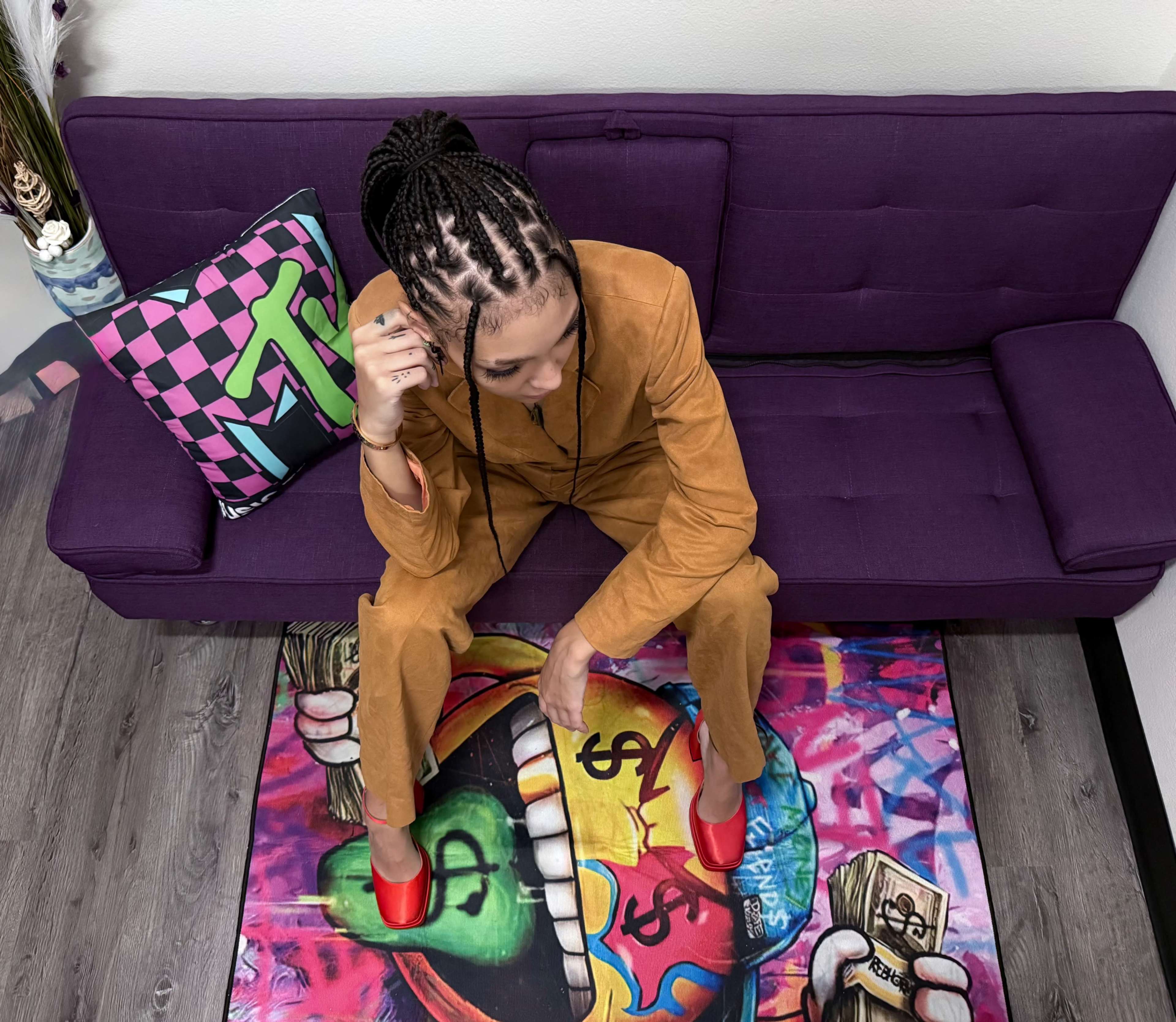 A person with braided hair sits on a colorful rug next to a purple couch, wearing a brown outfit and red shoes.