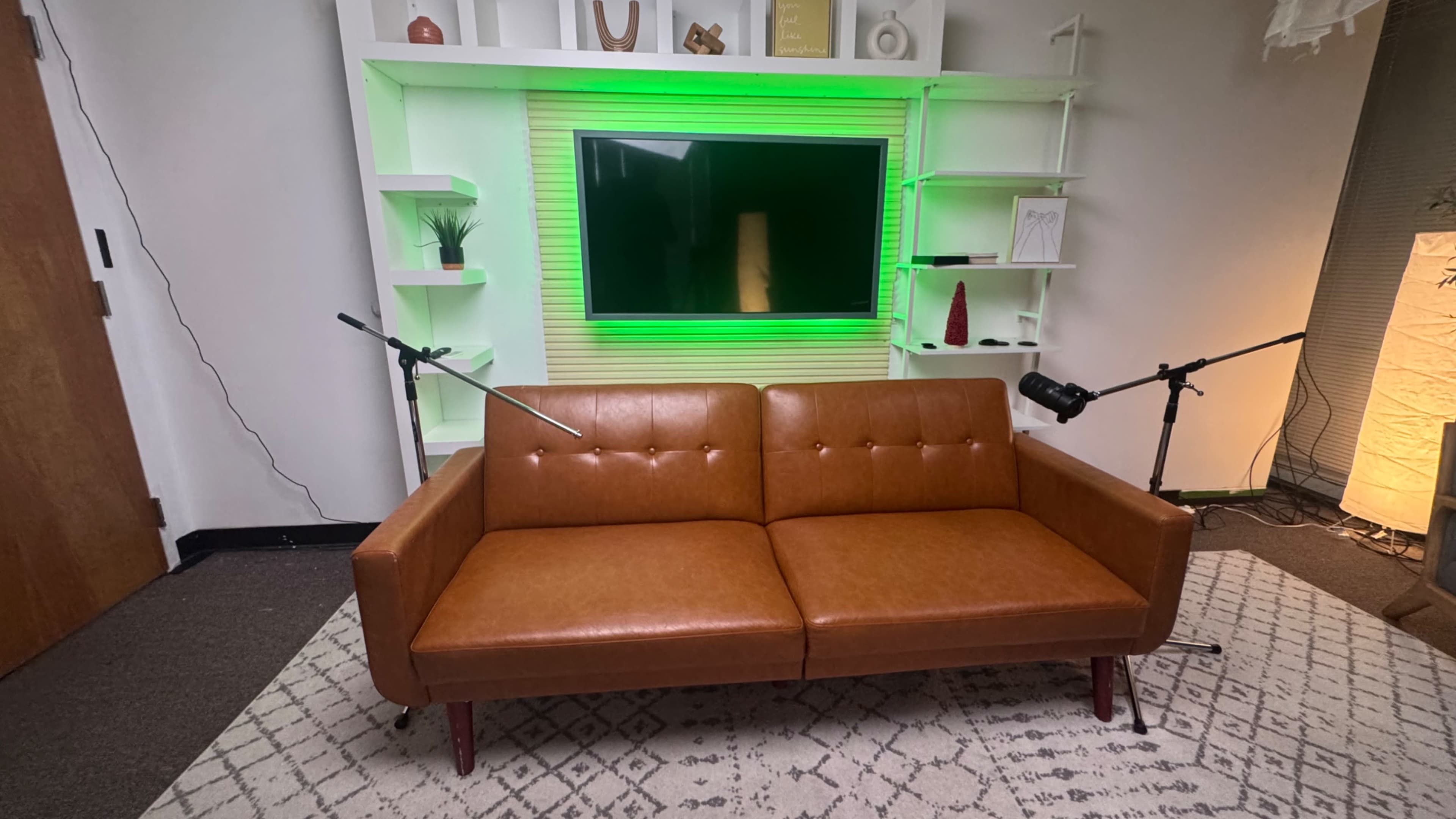 The image shows a modern brown leather sofa facing a wall-mounted television, with green backlighting and shelves on either side displaying decorative items.