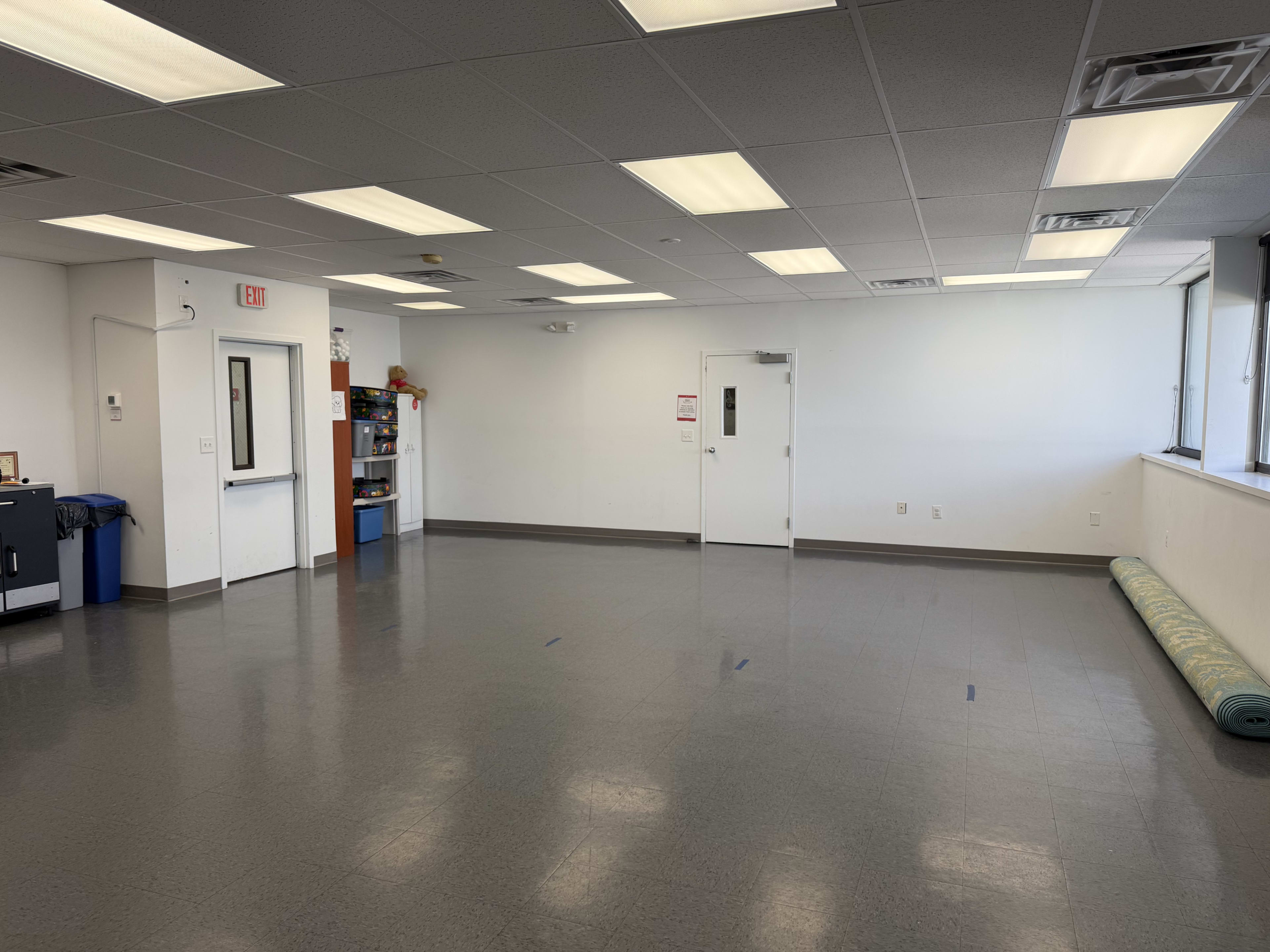 The image shows a spacious, empty room with gray flooring, white walls, and a single door on one side, along with a roll of green carpet positioned against the wall.