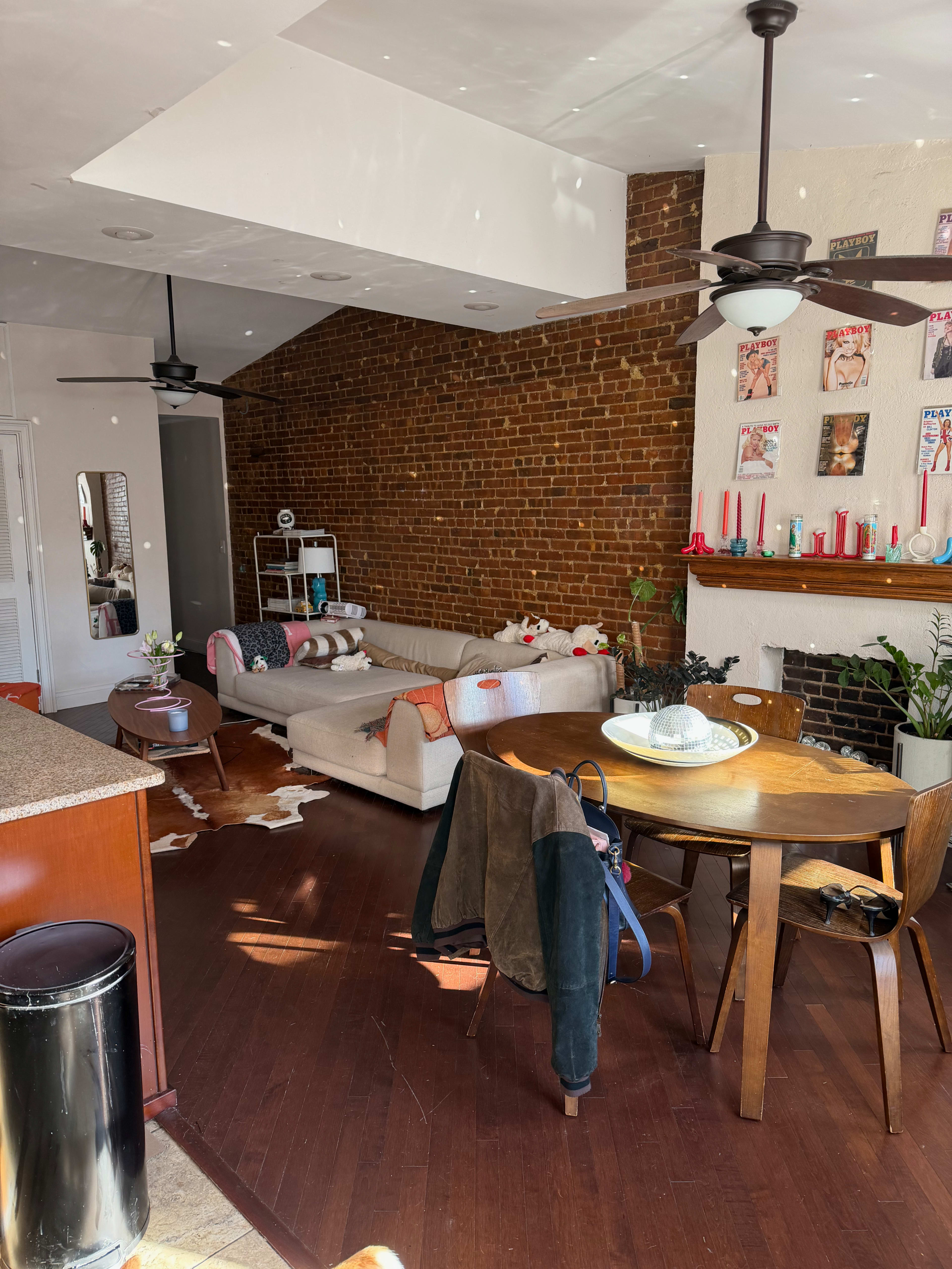 The image shows a modern living room with a brick wall, a couch, a dining table, a wooden floor, and decorative items on the walls and mantle.