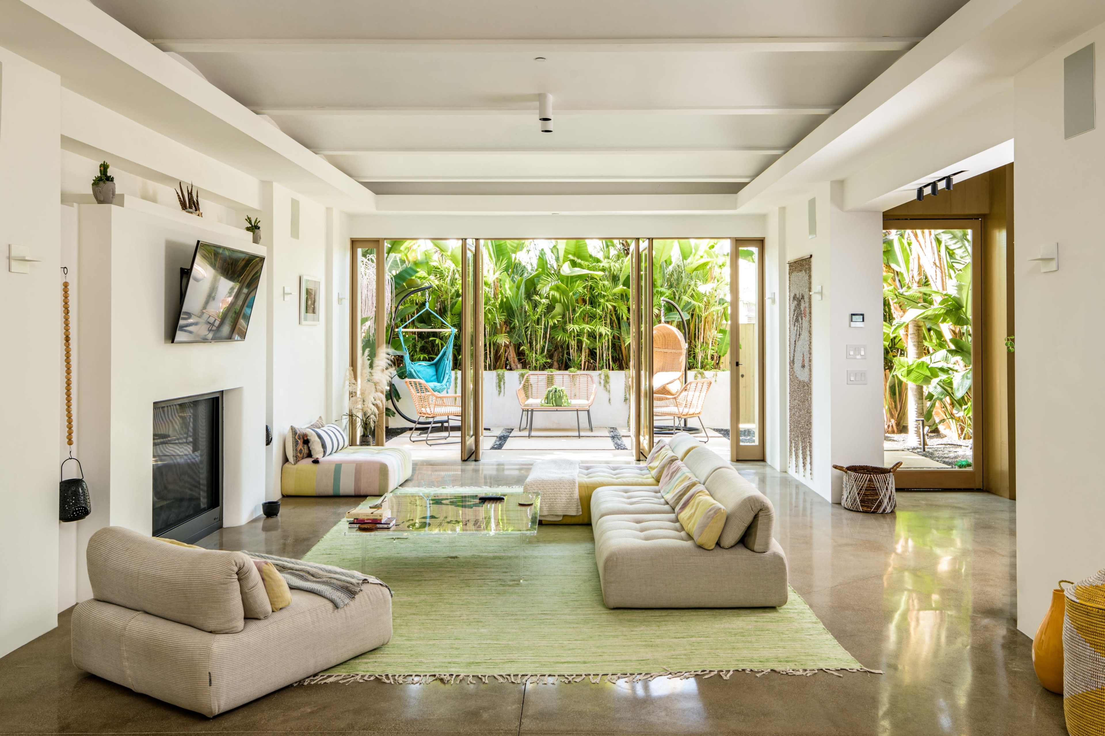 A spacious living room features a light-colored sectional sofa, a glass coffee table, and large sliding doors that open to a tropical backyard.