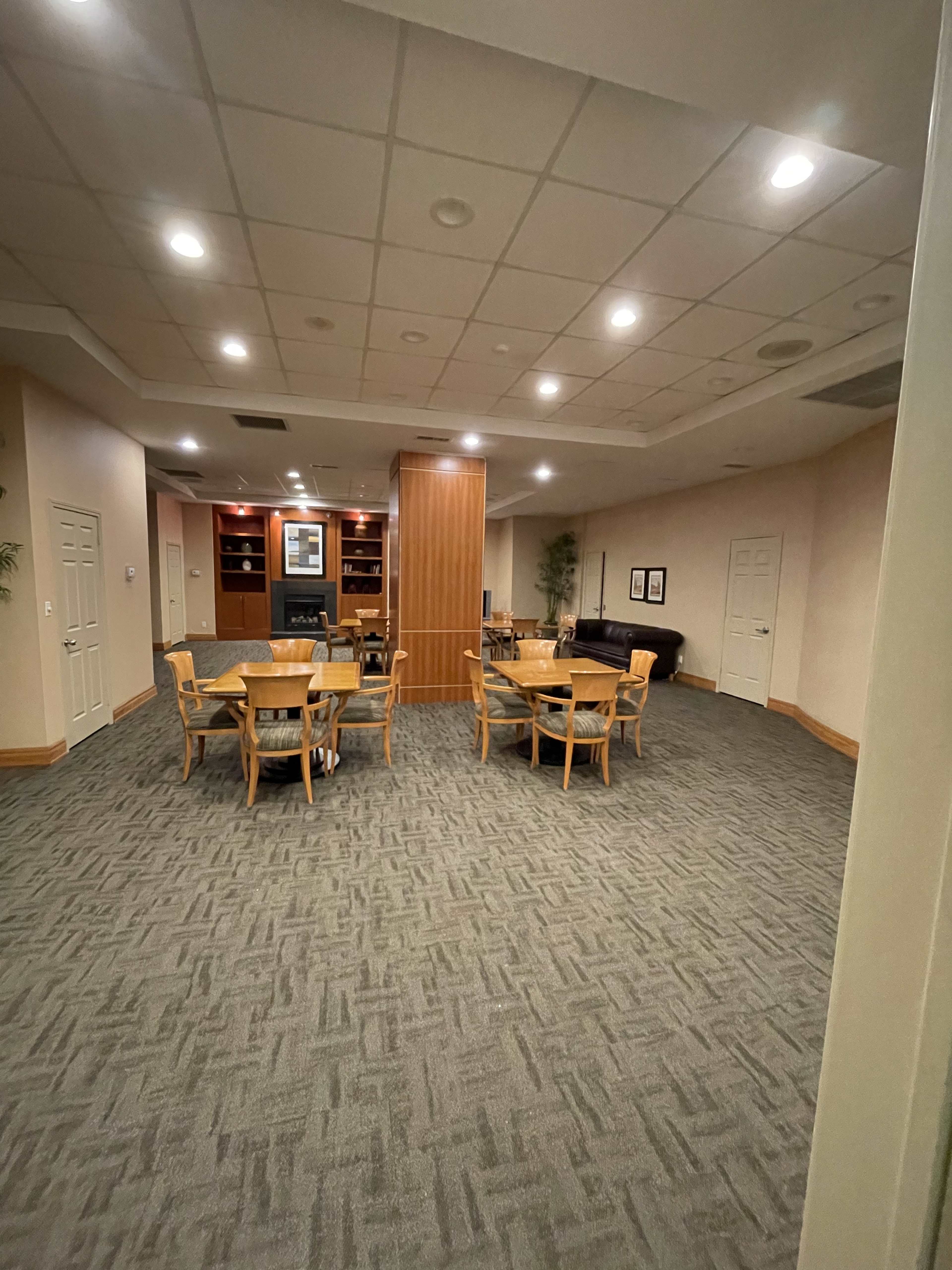 A spacious room with several wooden tables and chairs arranged around a central column, all set against a carpeted floor and soft lighting.