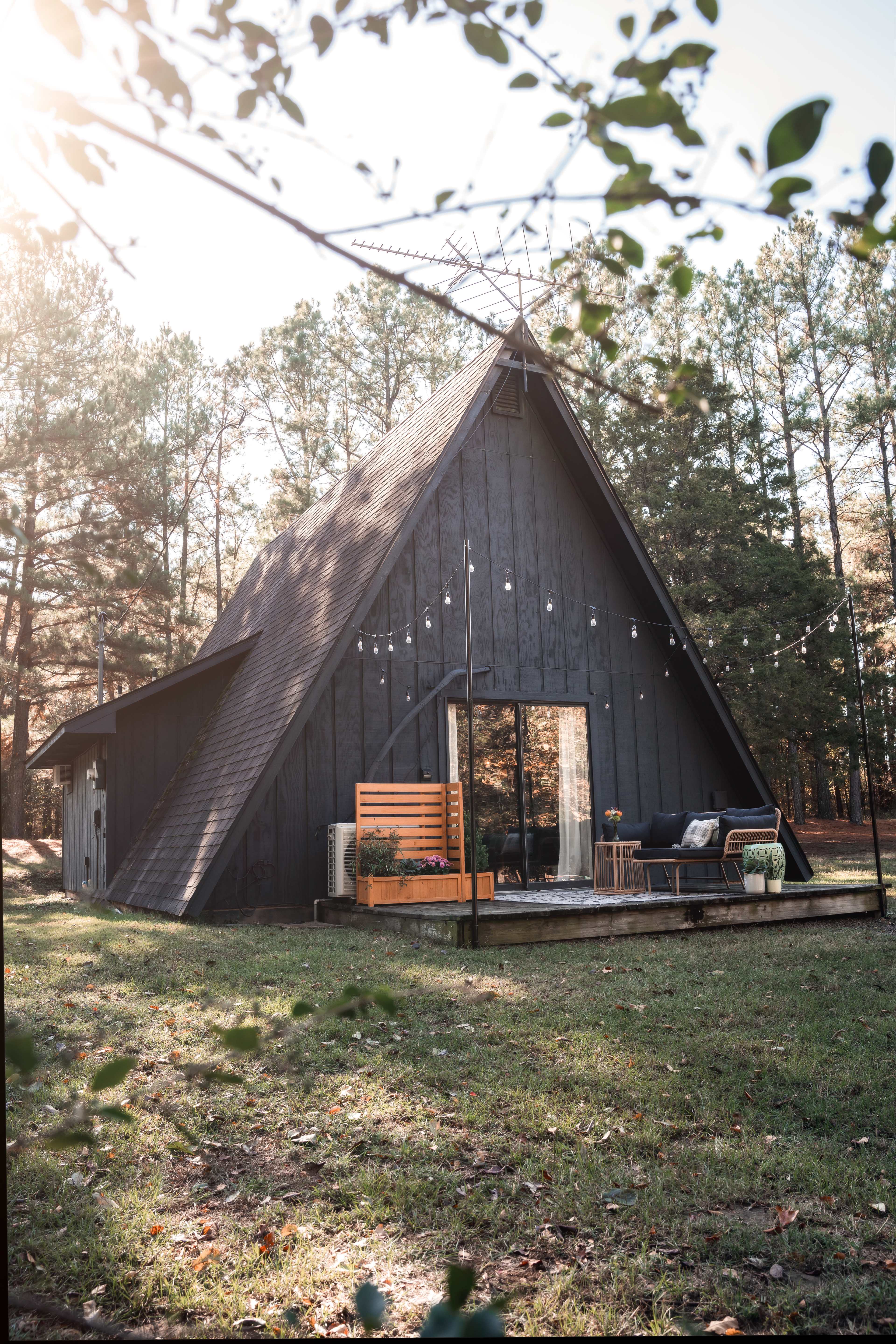 A black A-frame house surrounded by trees, featuring outdoor seating and decorative string lights.
