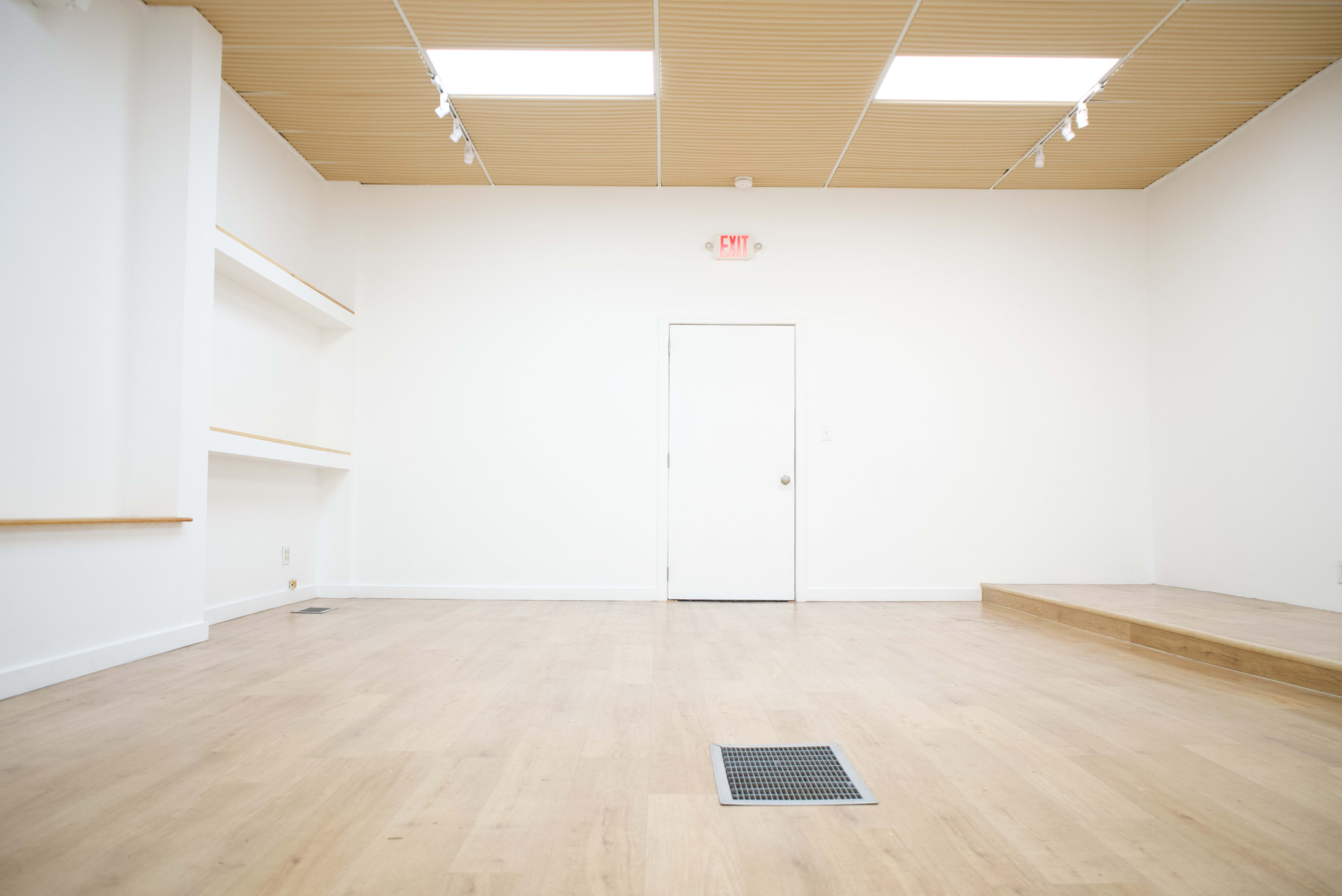 The image shows a minimalistic, empty room with light wooden flooring, white walls, and a closed door at the far end.