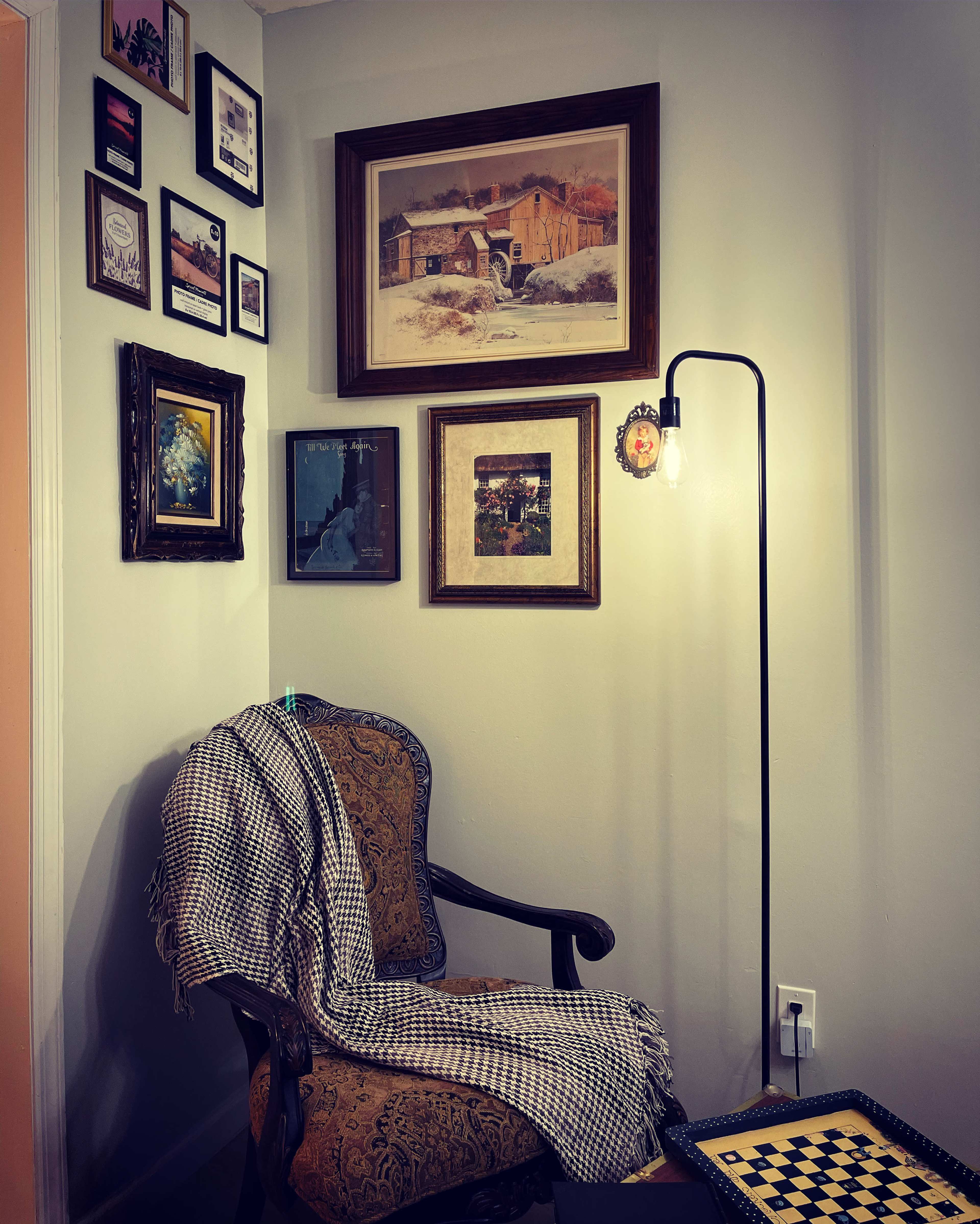 A corner of a room featuring an ornate chair draped with a blanket, a standing lamp, and an arrangement of framed artwork on the walls.