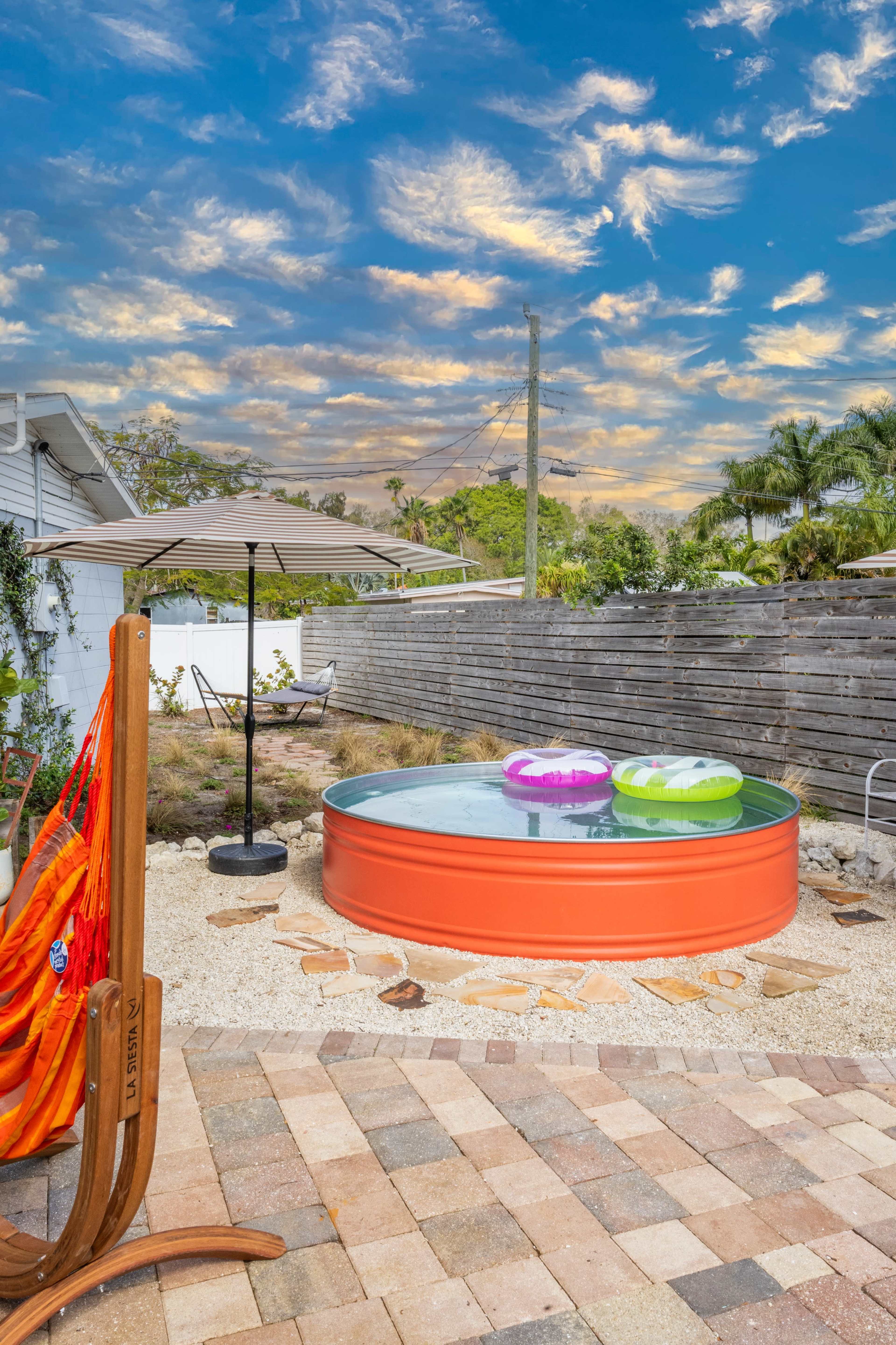 A circular metal pool with two floaties sits on a stone patio in a backyard with a hammock and an umbrella.