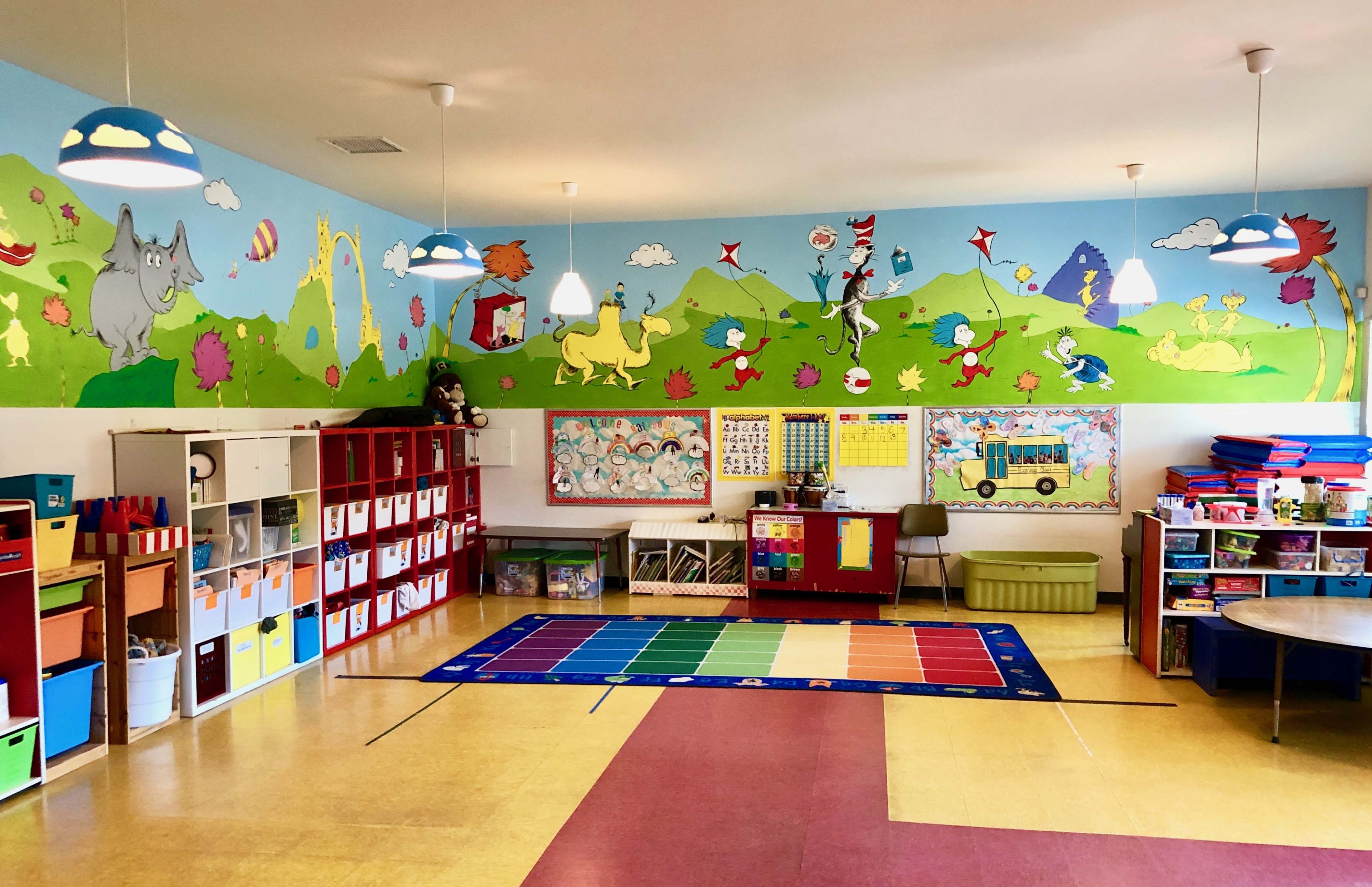 A bright and colorful kindergarten classroom with themed wall decorations, storage units, a large area rug, and educational materials displayed on the walls.