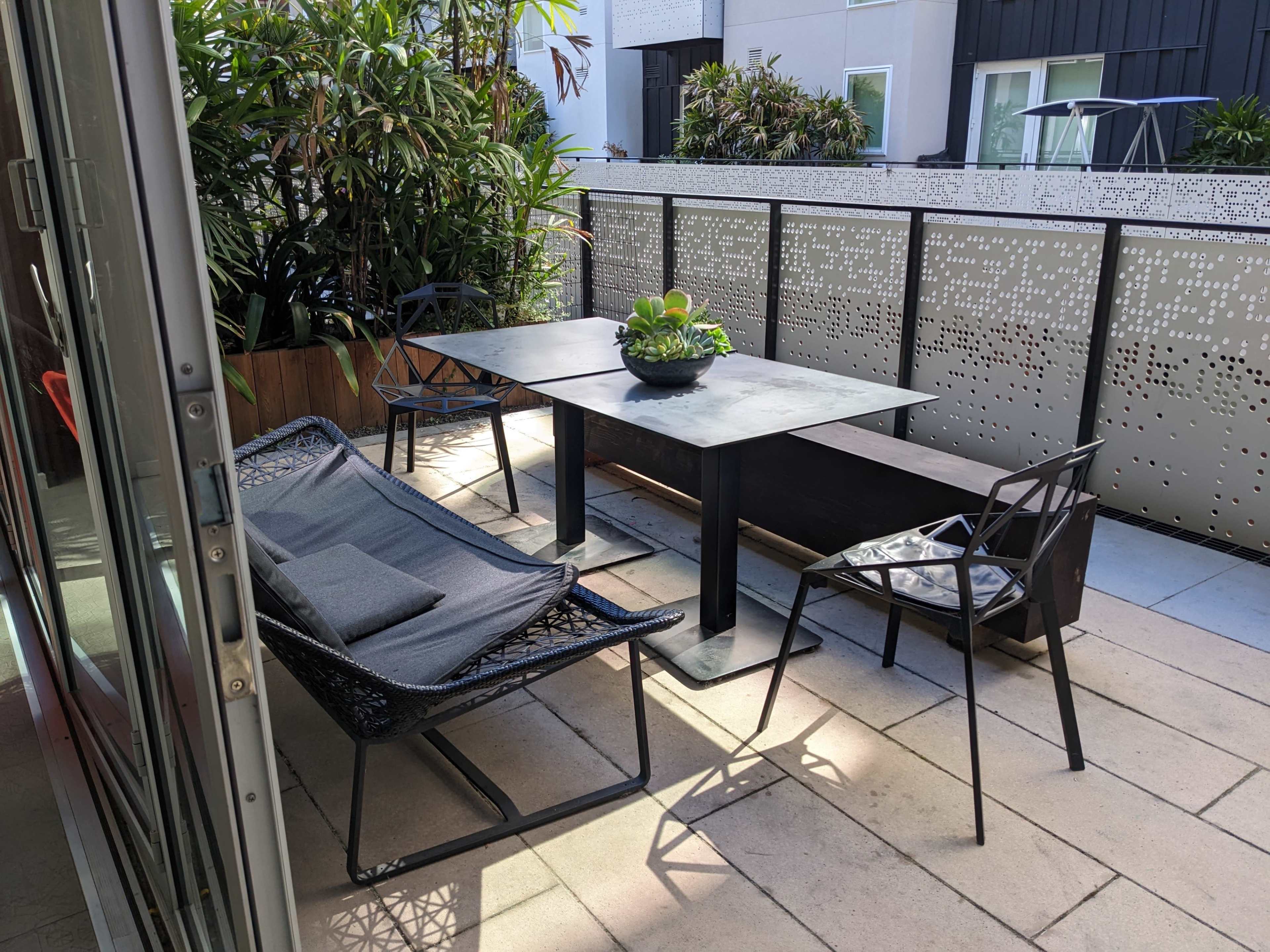 A patio area with a metal table, two chairs, and a bench, surrounded by lush plants and decorative partitioning.