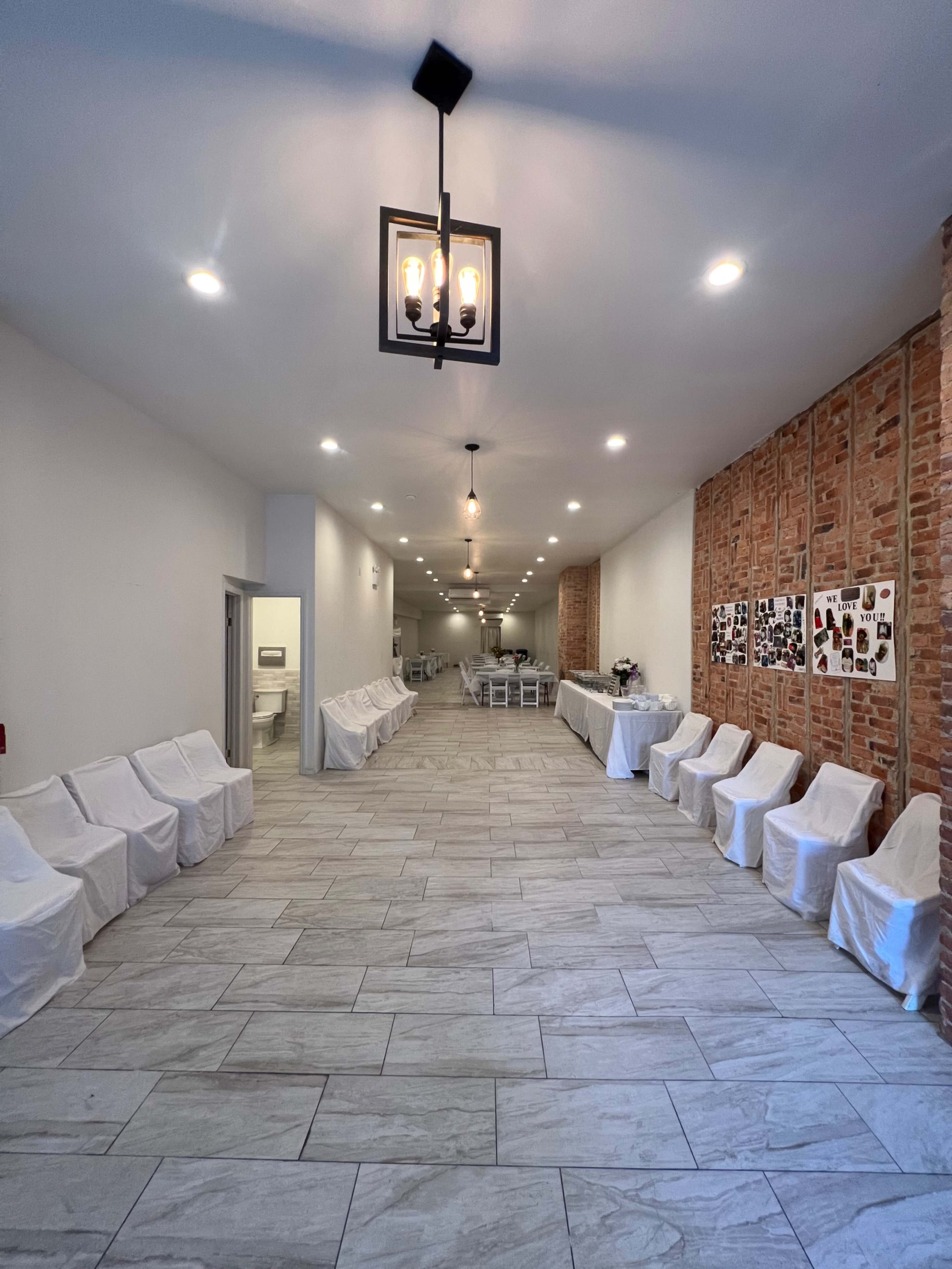 A long, narrow hall with covered chairs lined along each side and a brick wall featuring framed pictures.