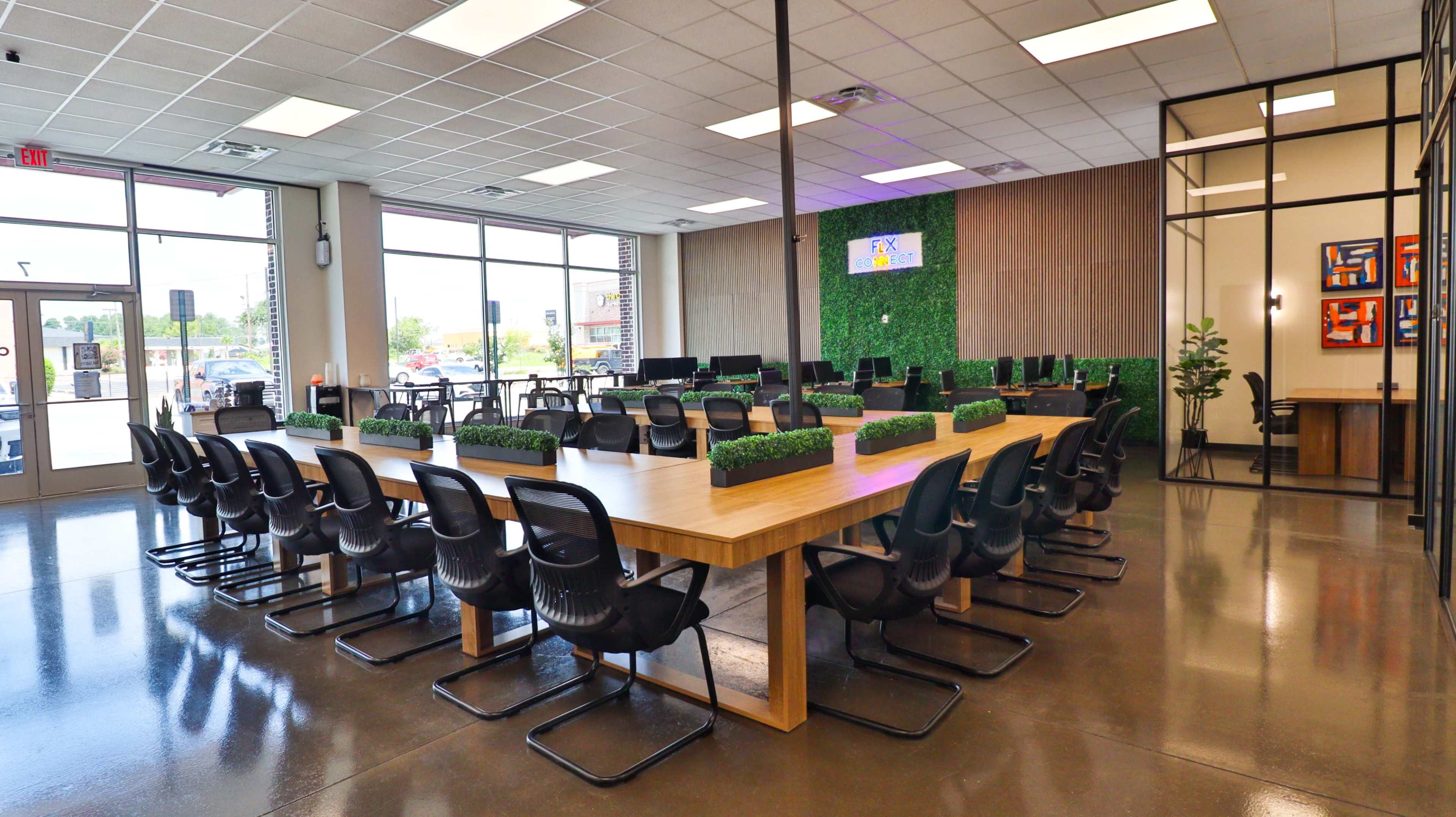 The image shows a modern office space with a large wooden table surrounded by black chairs, green potted plants, and multiple computer workstations in a well-lit environment.