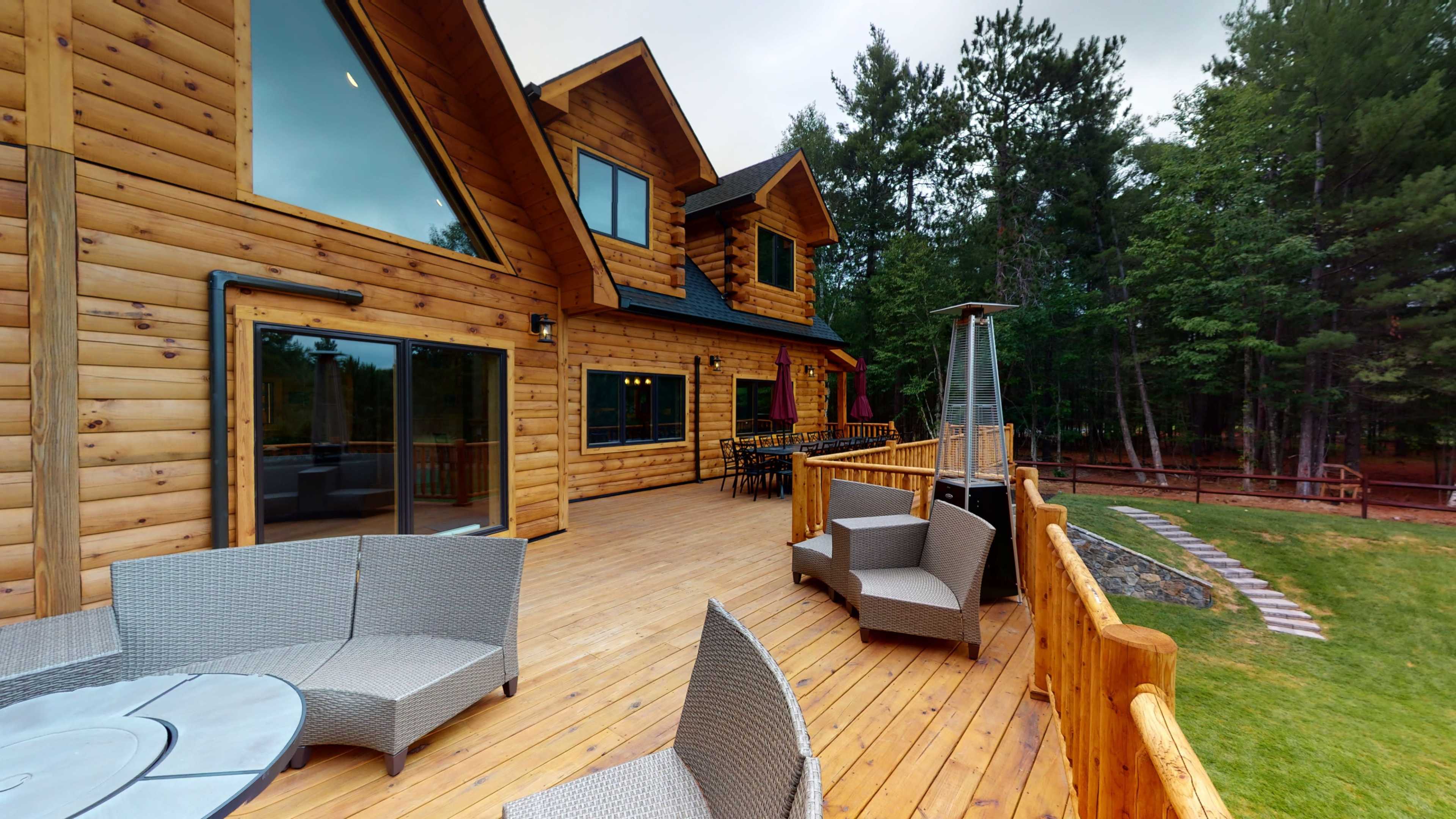 The image shows a wooden cabin with large windows and a spacious deck furnished with outdoor seating, surrounded by trees.