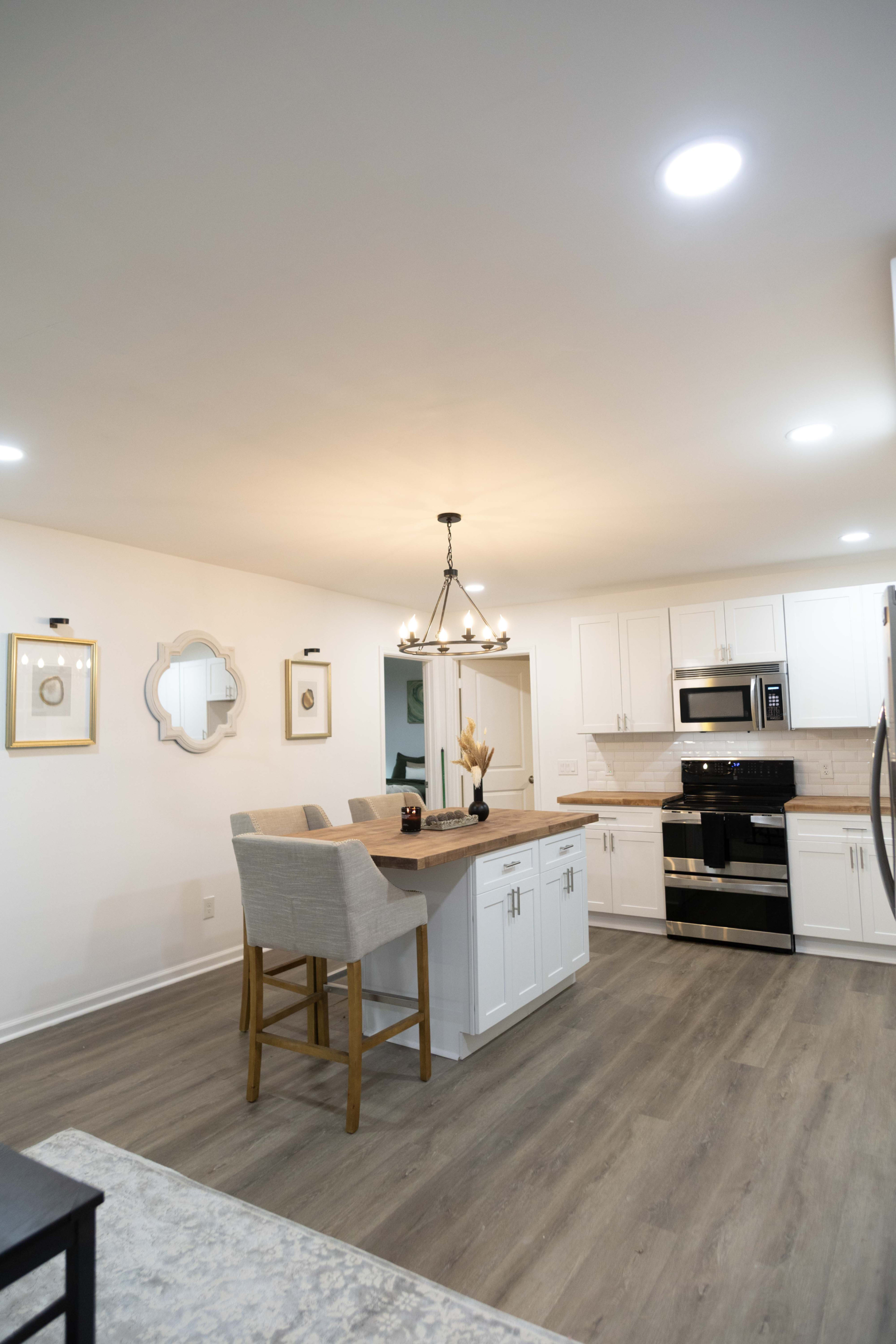 The image shows a modern kitchen featuring a central island with seating, white cabinets, stainless steel appliances, and bright overhead lighting.