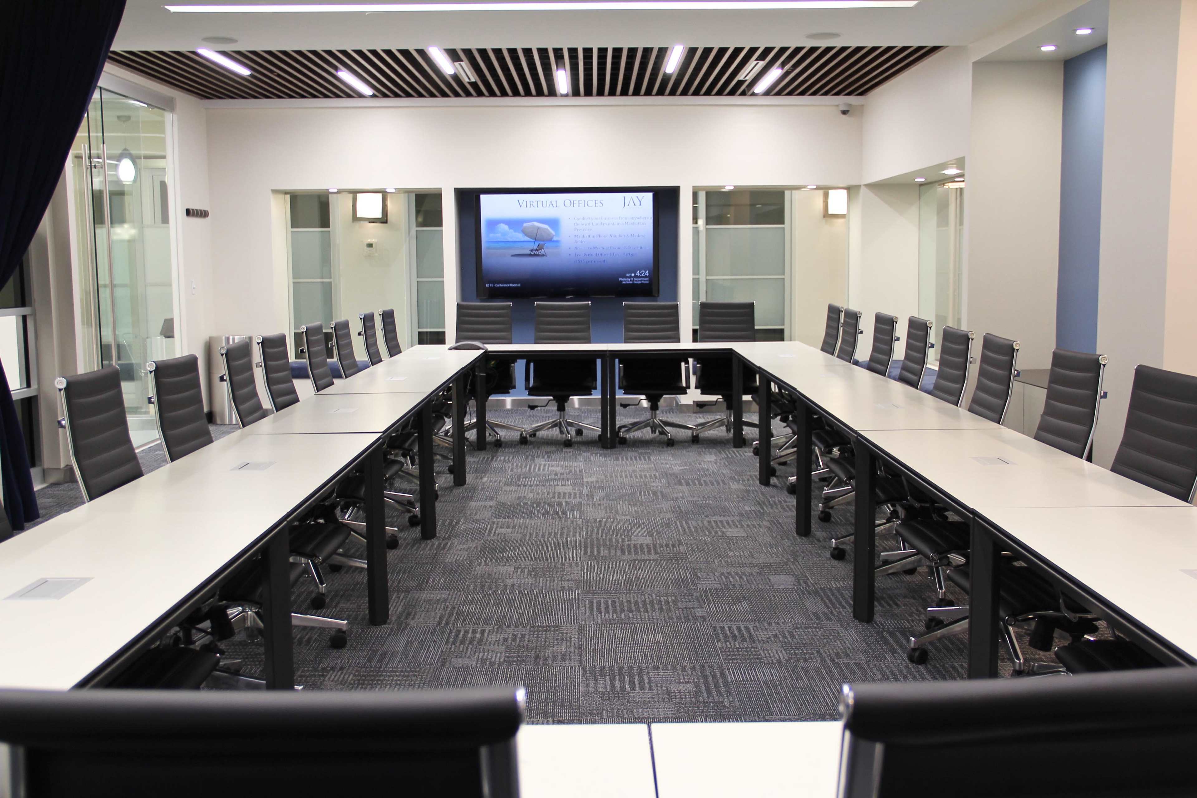 A spacious conference room features a large rectangular table surrounded by ergonomic chairs, with a large screen mounted on the wall displaying a presentation.