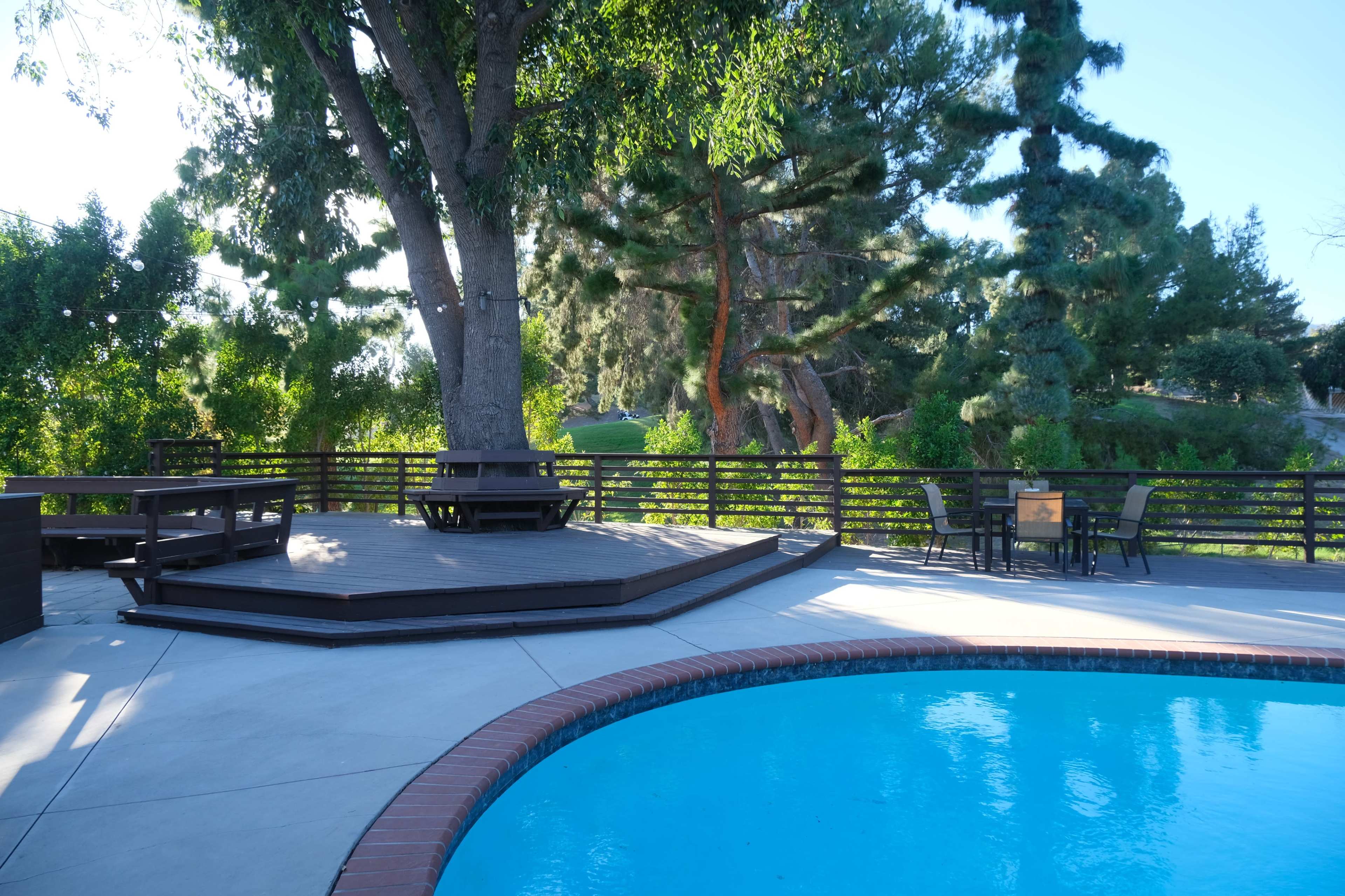 The image shows a backyard with a swimming pool, wooden deck, and seating areas surrounded by lush greenery.
