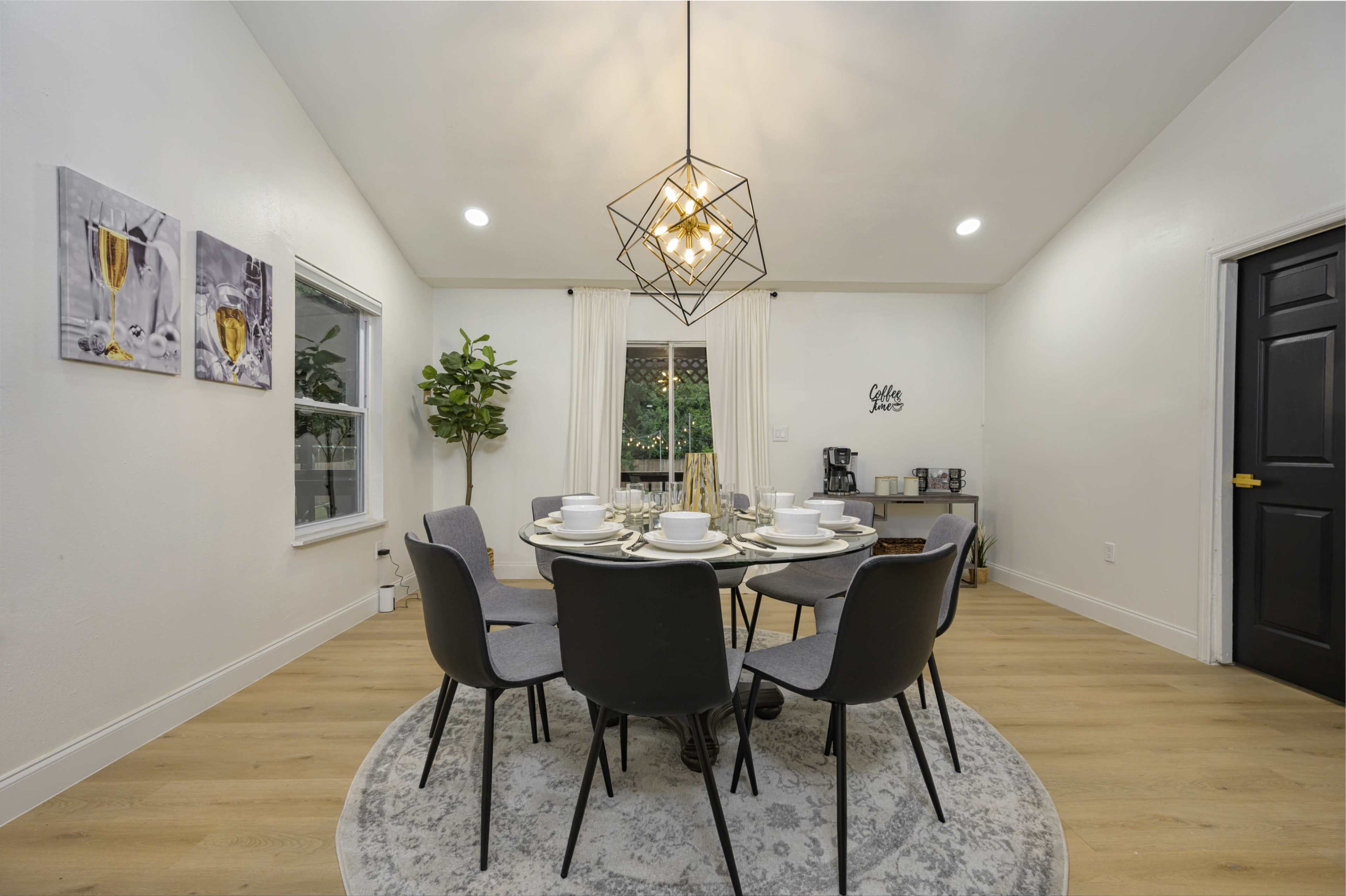 A dining area features a round table surrounded by six chairs, illuminated by a geometric pendant light, with two framed pictures on the wall and a coffee station in the corner.