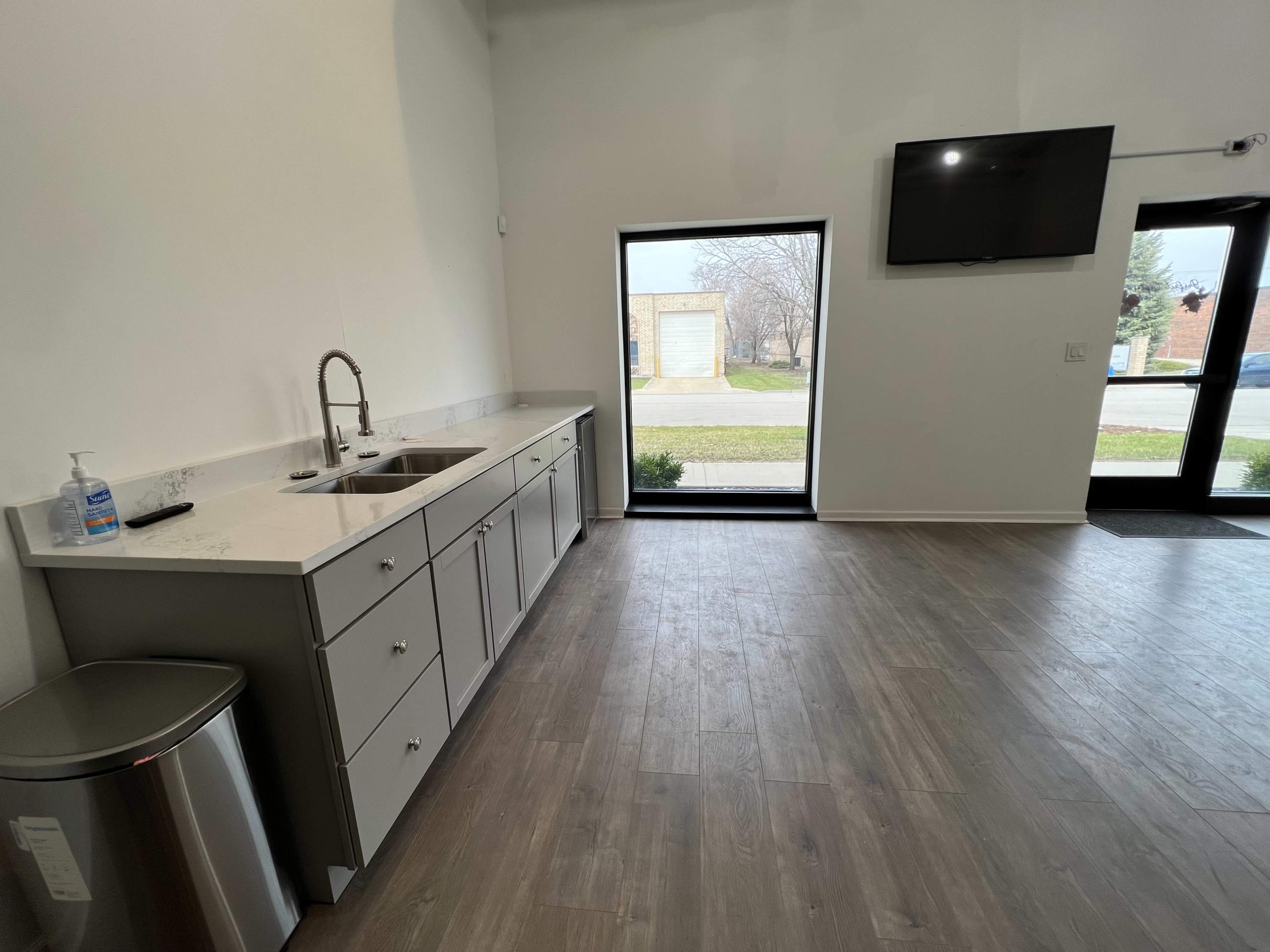 A modern kitchen space with a double sink, gray cabinetry, a television mounted on the wall, and a large window facing the outdoors.