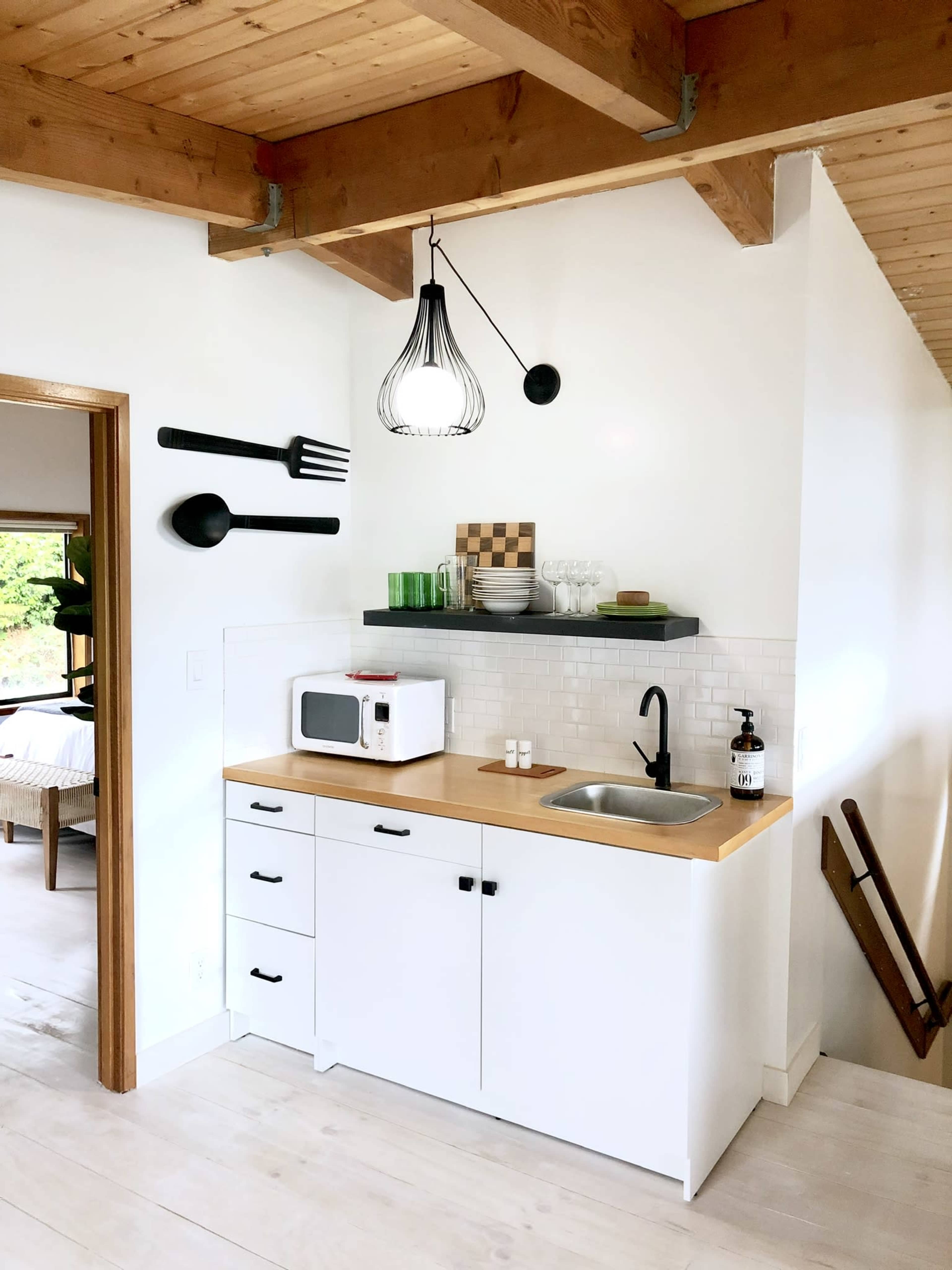 A compact kitchen features a white cabinet with a wooden countertop, a microwave, and a sink, complemented by decorative utensils on the wall and a black pendant light above.