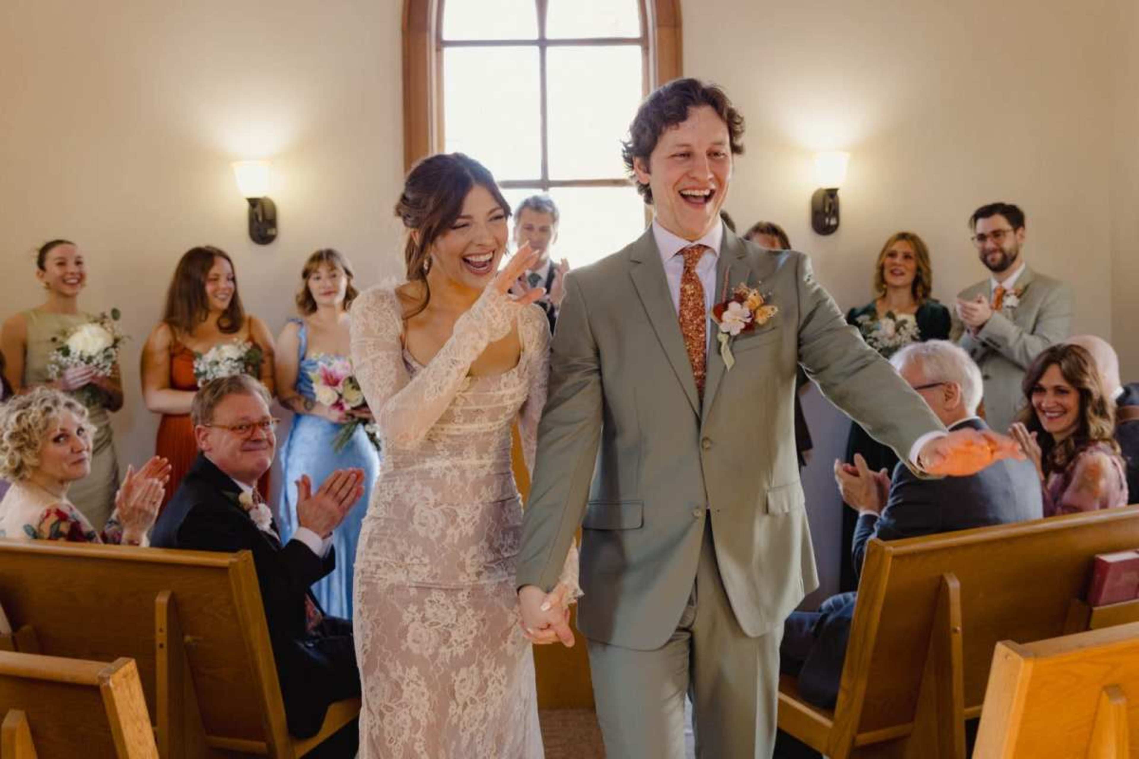 A bride and groom are joyfully walking down the aisle in a chapel, while guests applaud and celebrate their marriage.
