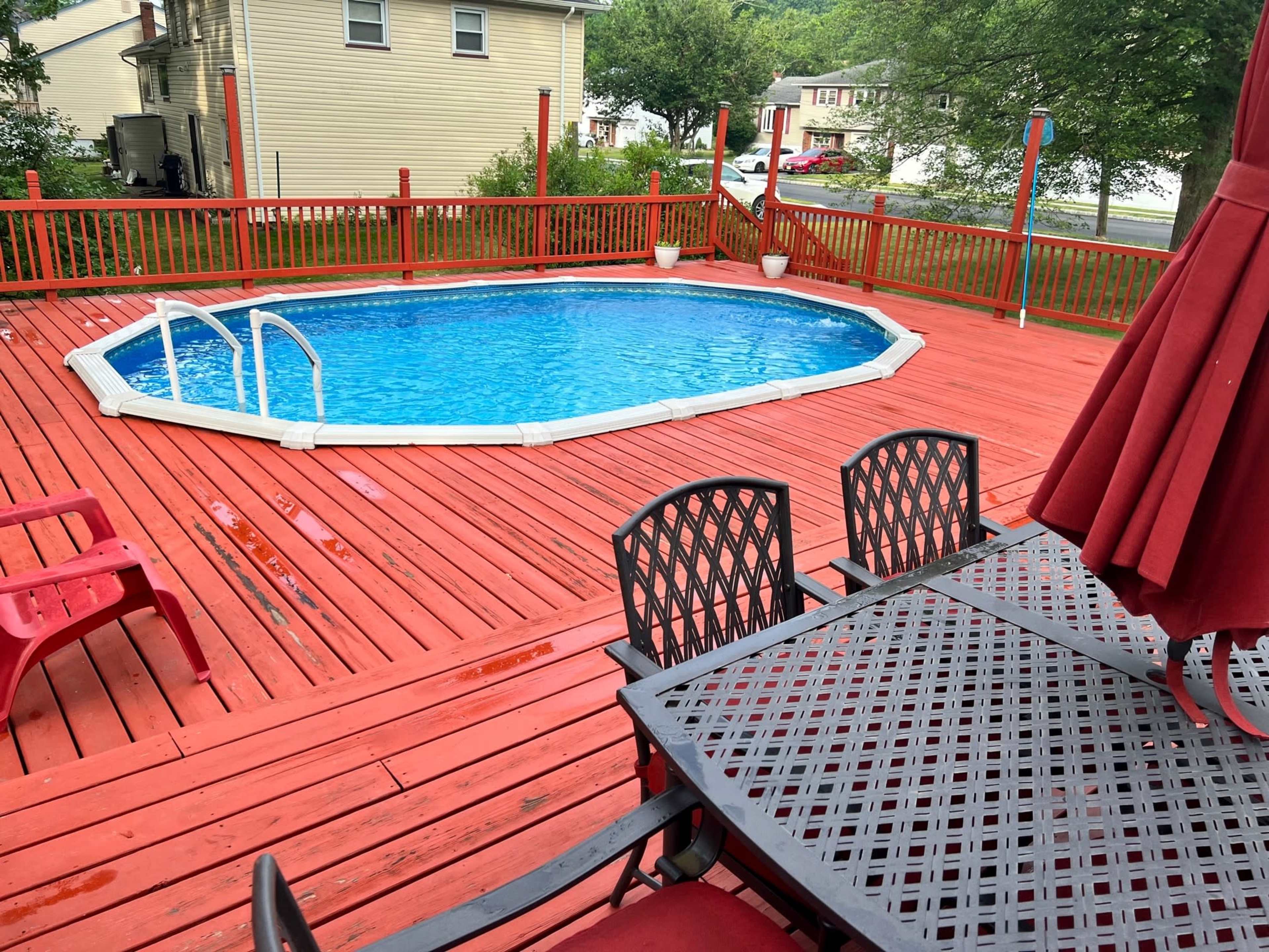 The image shows a red wooden deck with a blue oval pool surrounded by chairs and tables.