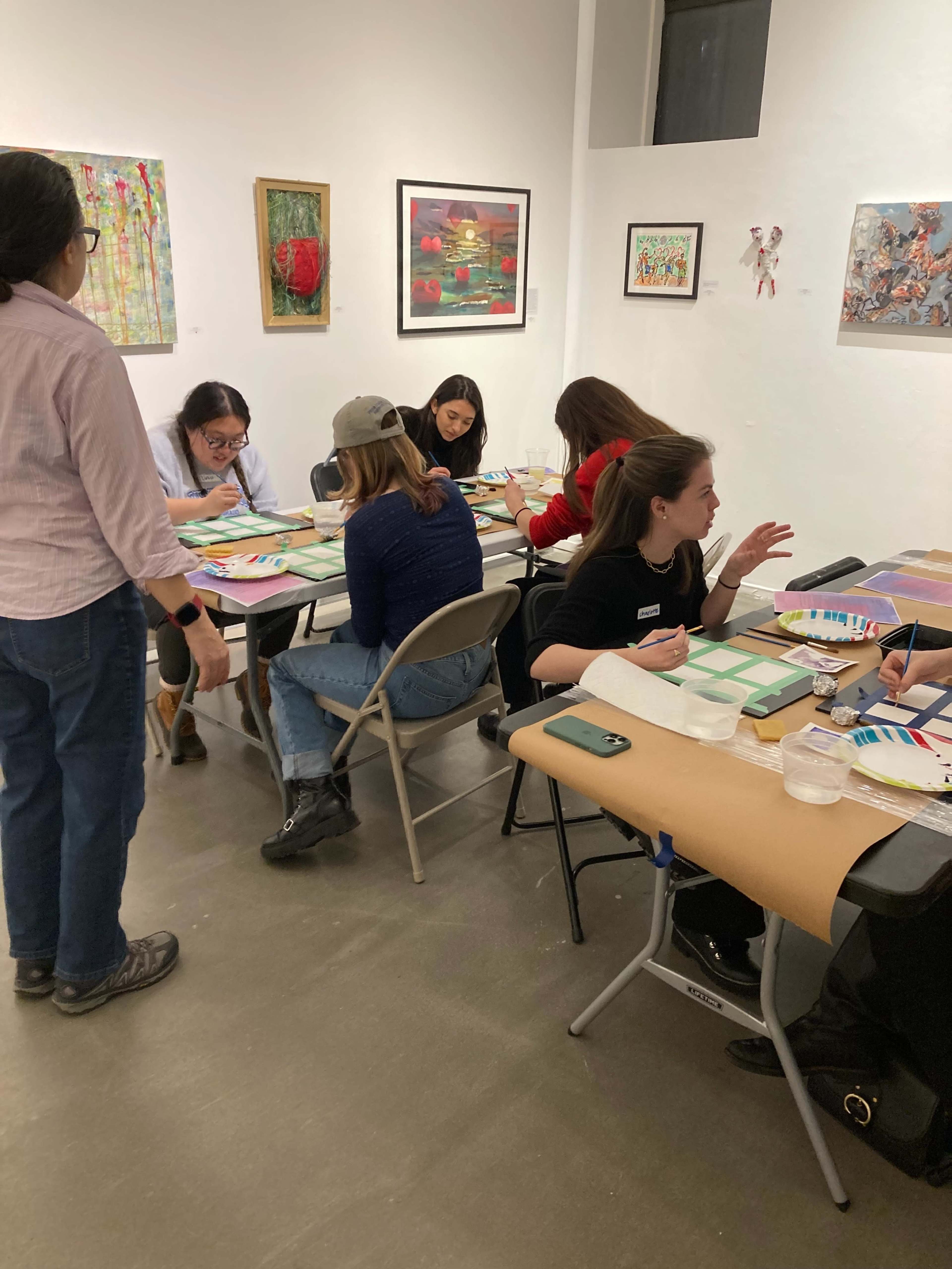 A group of people sits at tables in an art studio, engaged in creating artwork while surrounded by various paintings on the walls.